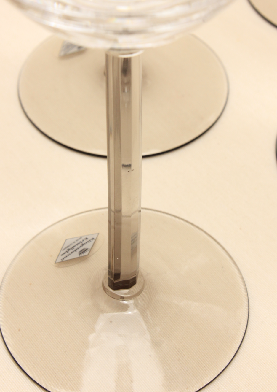 A close-up shows a Crystal Wine Glass from Boga Avante Shop, featuring a transparent design with a thin vertical stem and circular base. Set on a beige surface, it boasts contemporary elegance highlighted by a small handcrafted label on the base.