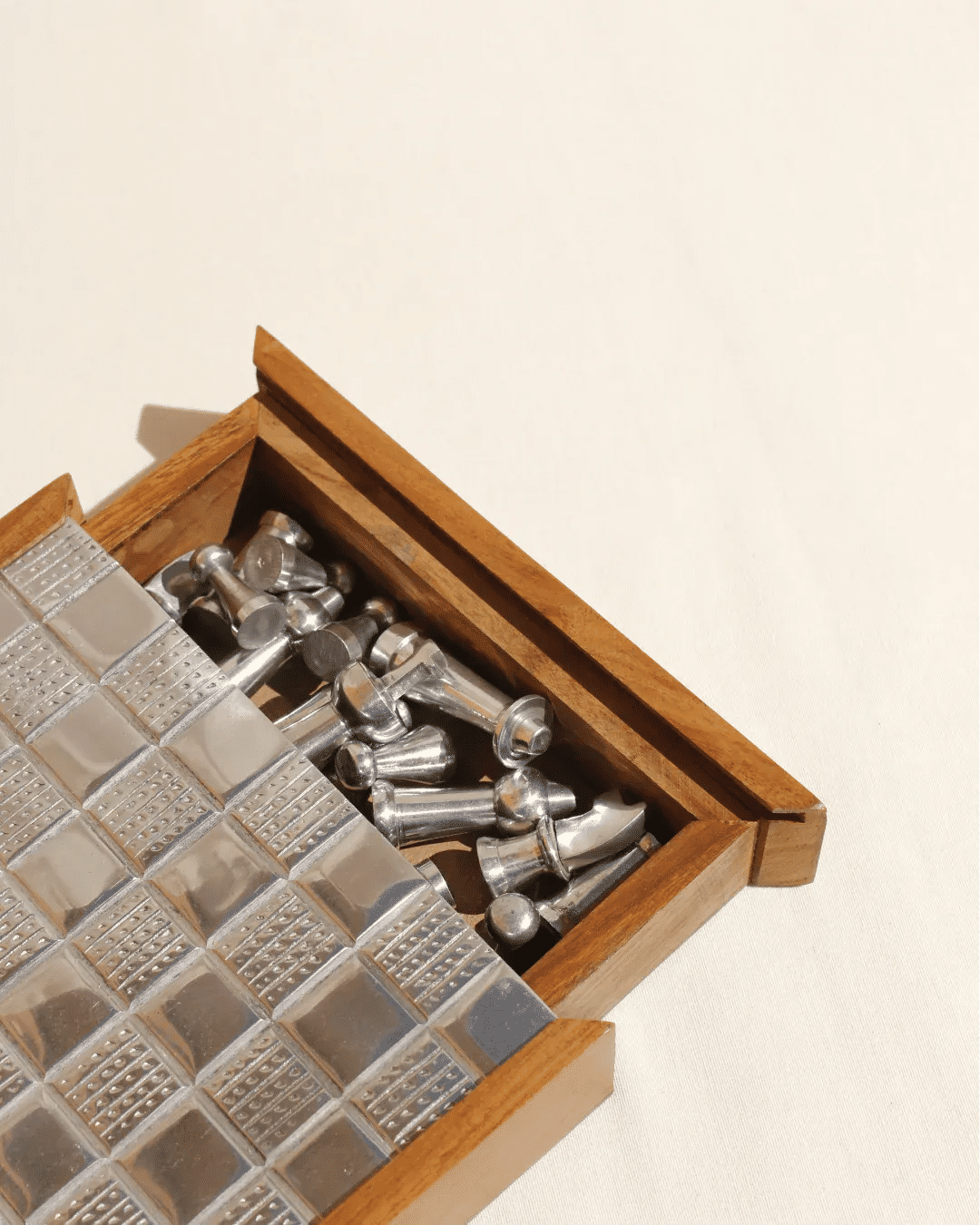 The Wood and Metal Chessboard by Boga Avante Shop features a wooden box with an array of shiny tiles and metallic chess pieces inside, positioned on a cream surface to enhance any luxury interior with contemporary design.
