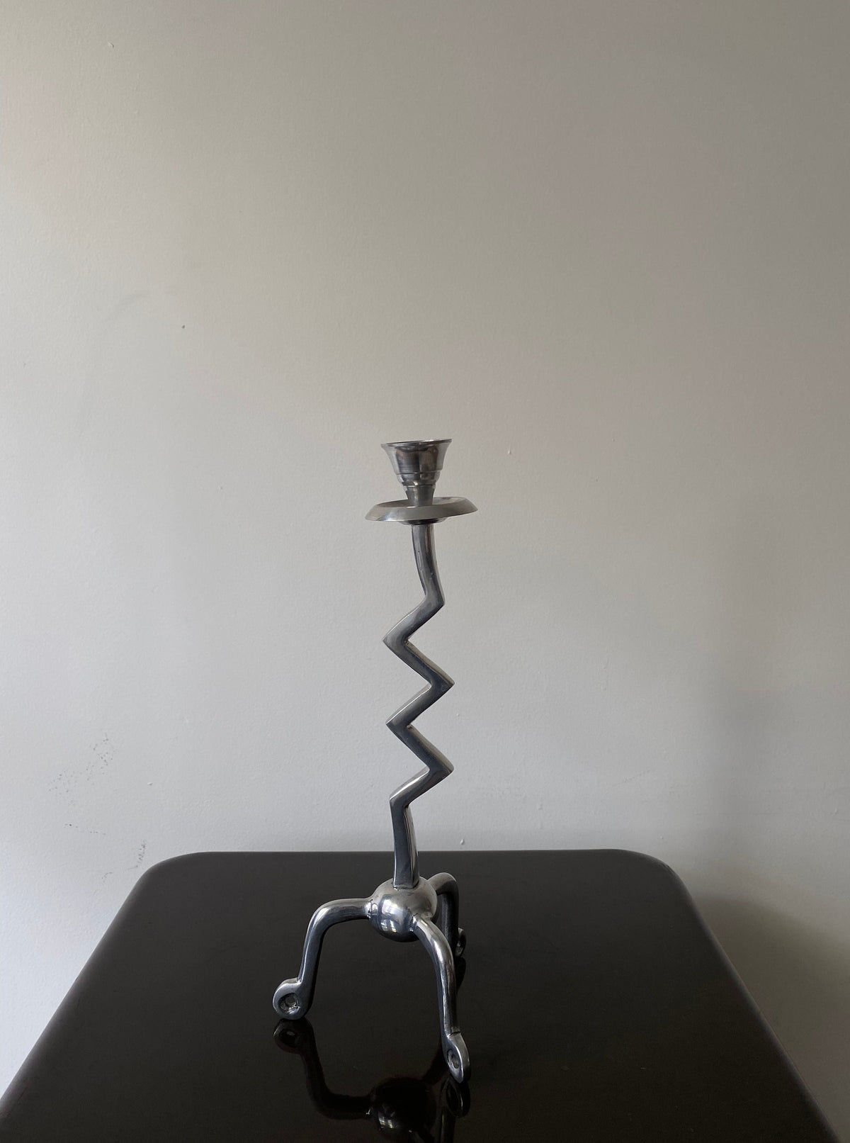 The Vintage Zigzag Candle Holder by Les Objets Amsterdam is a luxury interior piece with a minimalist design. It features a zigzag stem, three curved legs, and stands elegantly on a glossy black surface against a plain white wall.