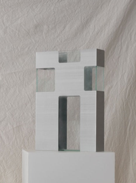 A modern sculpture featuring a minimalist design of metal rectangles and squares interlocked with plexiglass panels, displayed against a crinkled white fabric background by Les Objets Amsterdam.