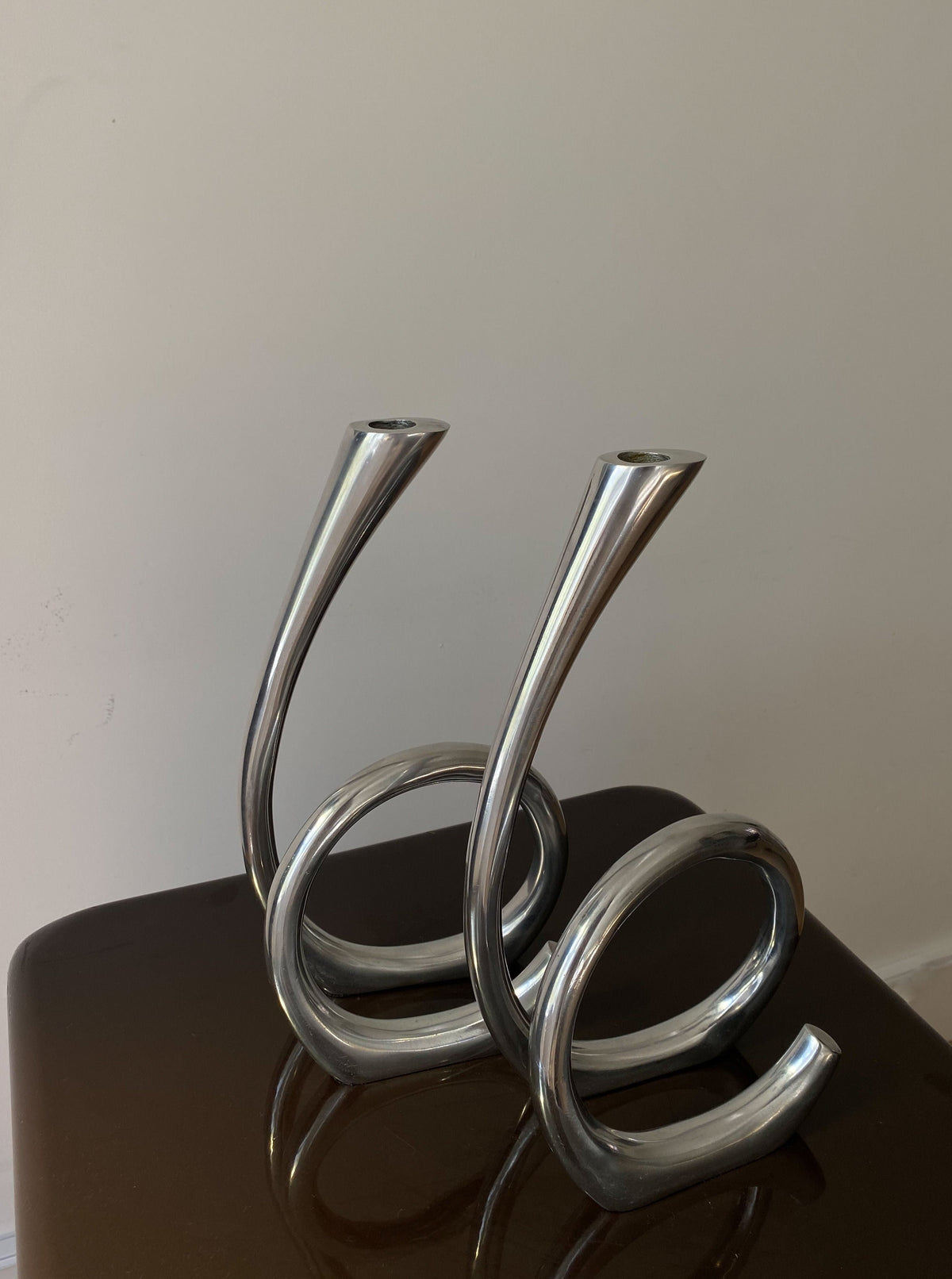 Two modern, shiny silver Vintage Curly RVS candle holders with a twisted metal design, displayed on a dark reflective surface against a light-colored wall by Les Objets Amsterdam.