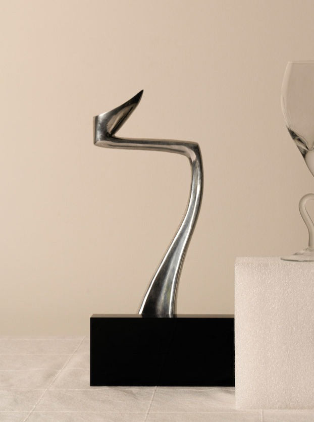 A modern abstract sculpture made of shiny silver metal, featuring fluid curves and angles, is displayed on a black rectangular base—a true testament to timeless design. To the right, part of a transparent glass sits on a white textured surface. A beige wall serves as the backdrop. The sculpture in question is the Matthew Hilton Swan - Medium by Les Objets Amsterdam.