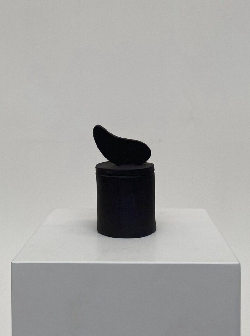 A minimalist Sculptural Scented Candle No. 35 of a black abstract shape positioned atop a cylindrical black base, displayed on a white pedestal against a light gray background by 2222Studio.