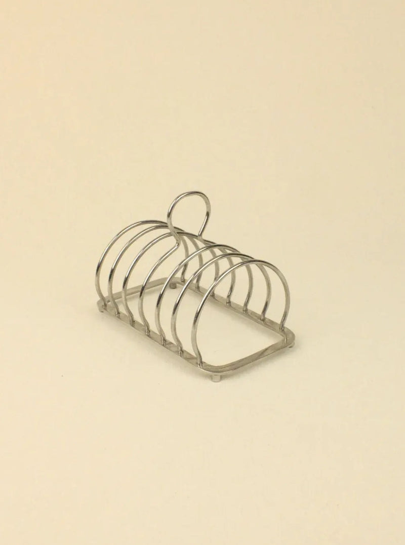 The Art Deco Toast Holder by Boga Avante Shop is a silver rack with five slots and a circular handle, set on a light beige background for a minimalist aesthetic.