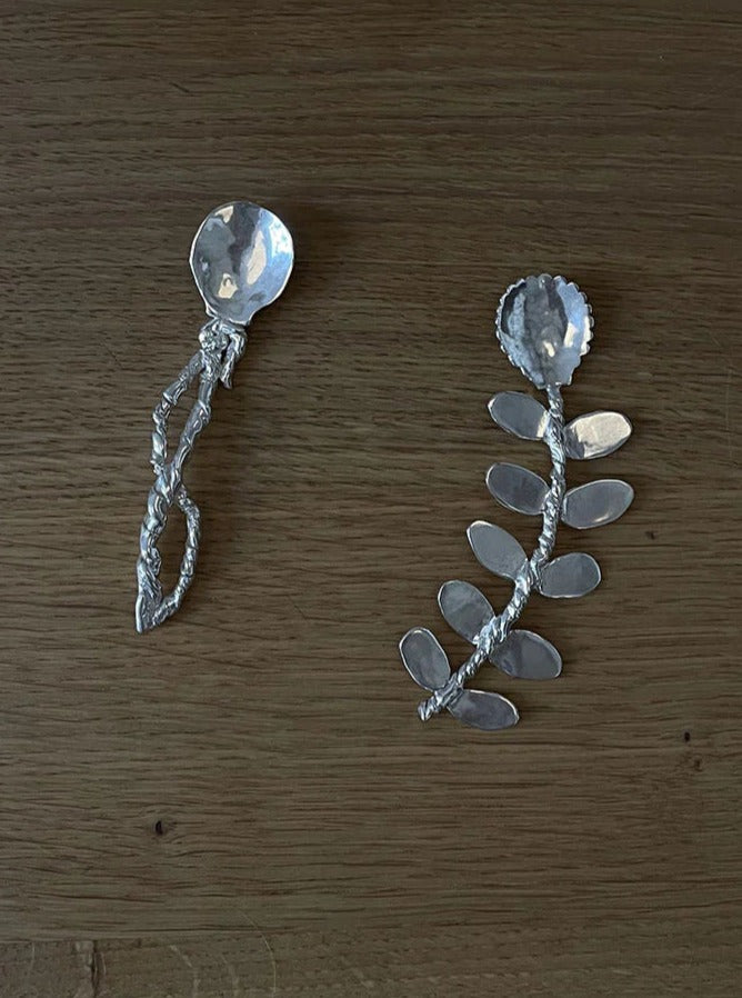 Two unique handcrafted Corali sterling silver Babette Compote Spoons with decorative handles on a wooden surface, one with a spiral design and the other with a branch and leaf motif.