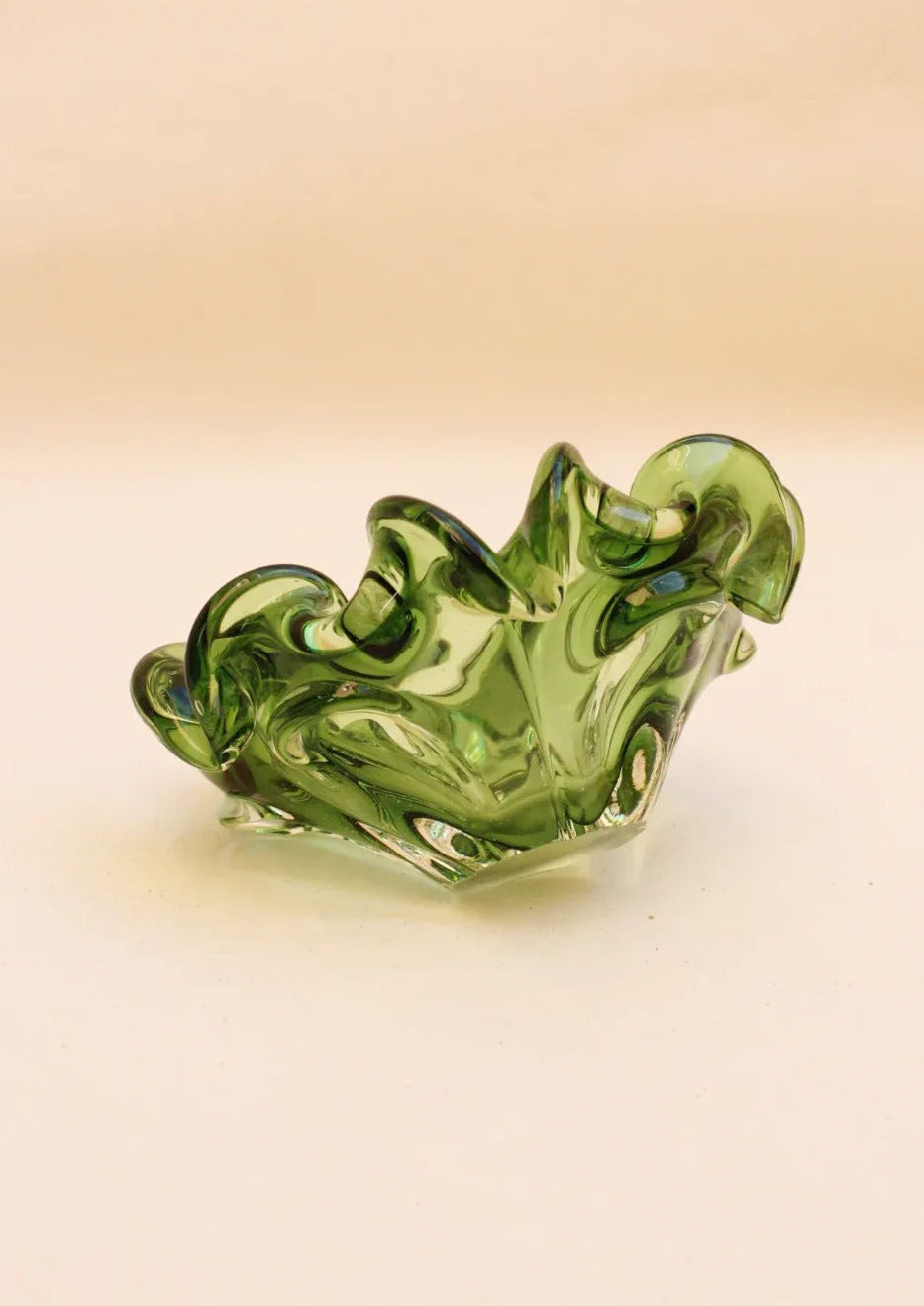 The Murano Glass Bowl by Boga Avante Shop is a decorative piece with a wavy, abstract design in translucent green. Its artistically curved and twisted edges offer contemporary flair, enhancing any luxury interior against a plain background.