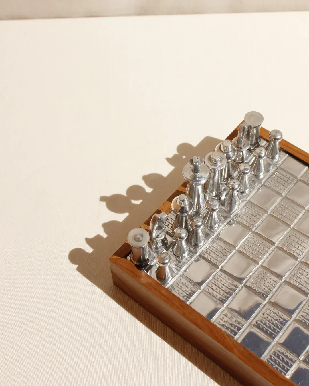 A Wood and Metal Chessboard from Boga Avante Shop features a glossy tiled surface, wooden frame, and metallic chess pieces. The minimalist design casts distinct shadows on the light-colored background.