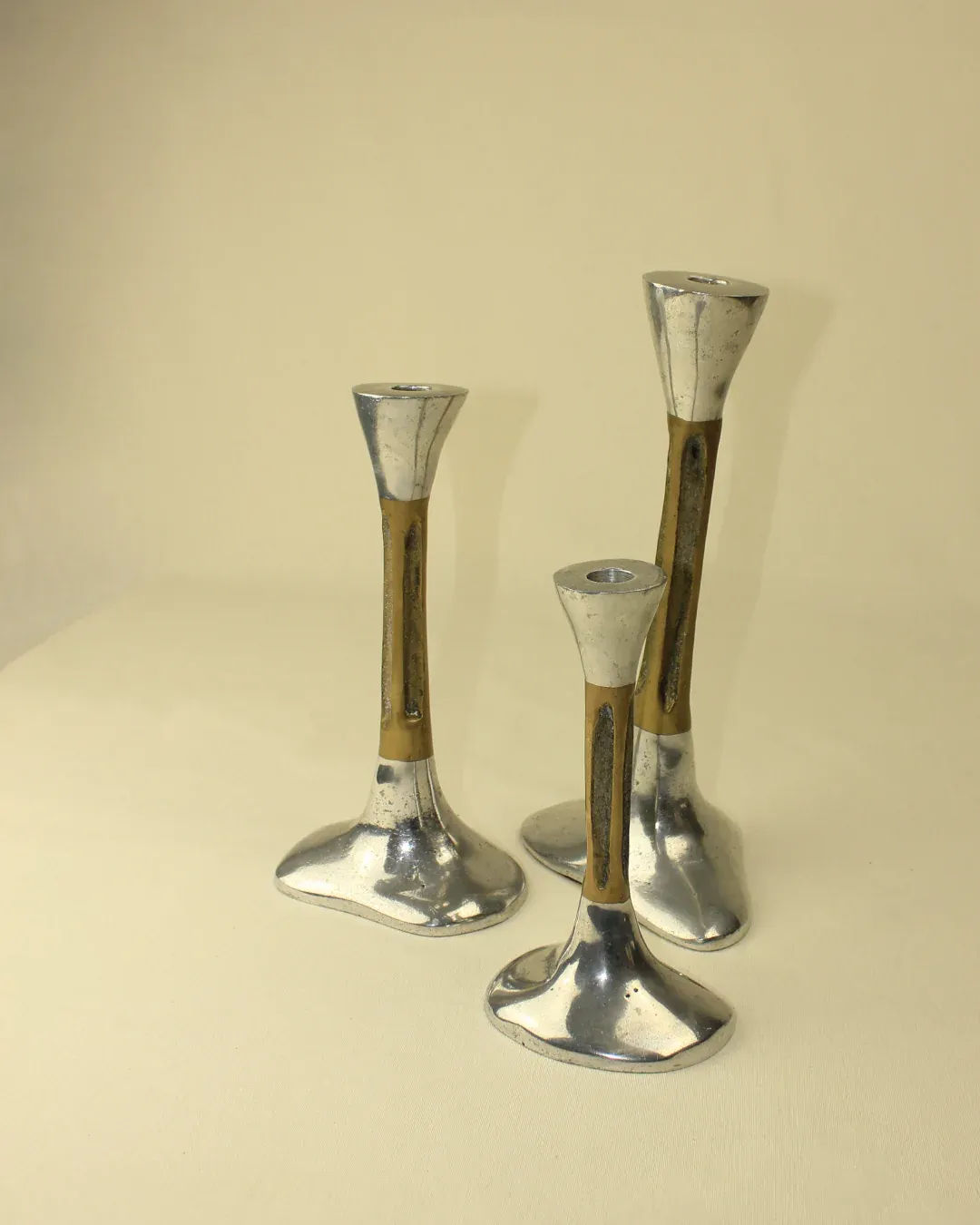 A Trio of Brutalist Candleholders by Boga Avante Shop are tapering metal pieces with a distinctive design, featuring a slim middle section and wide bases and tops. Displayed on a flat surface against a plain background, each candleholder varies slightly in height, exuding a modern, brutish design aesthetic perfect for home decor.