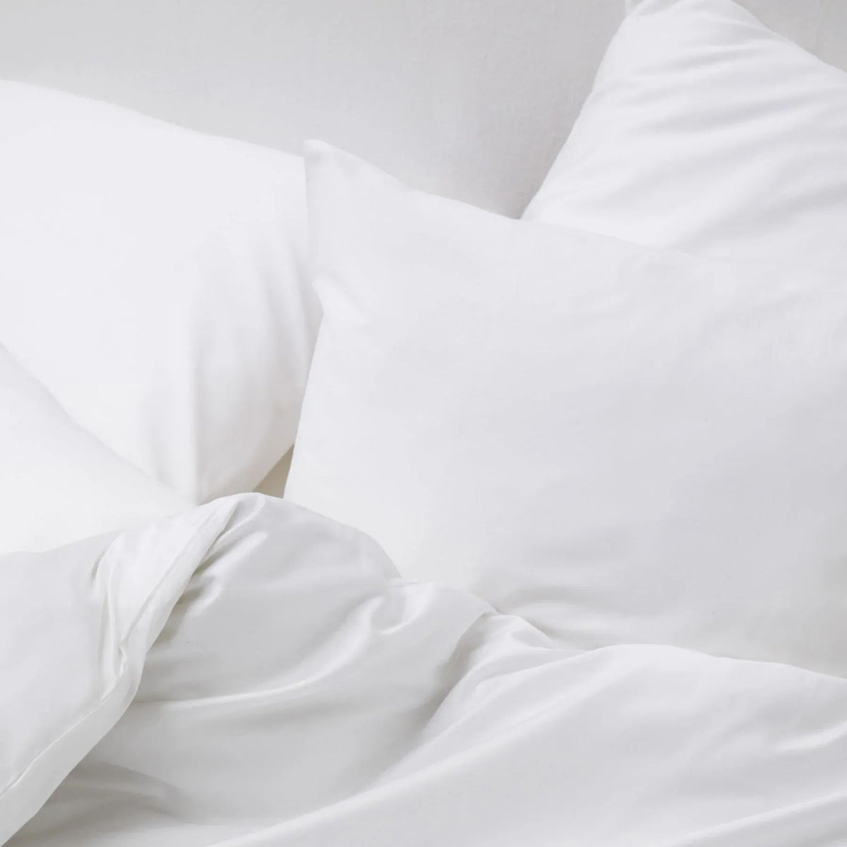 Close-up of the Cotton Sateen Bed Set in White by MOST, featuring two pillows and a rumpled blanket. The minimalist setting highlights the soft and inviting linens, exuding luxury and relaxation with their fresh, clean appearance.