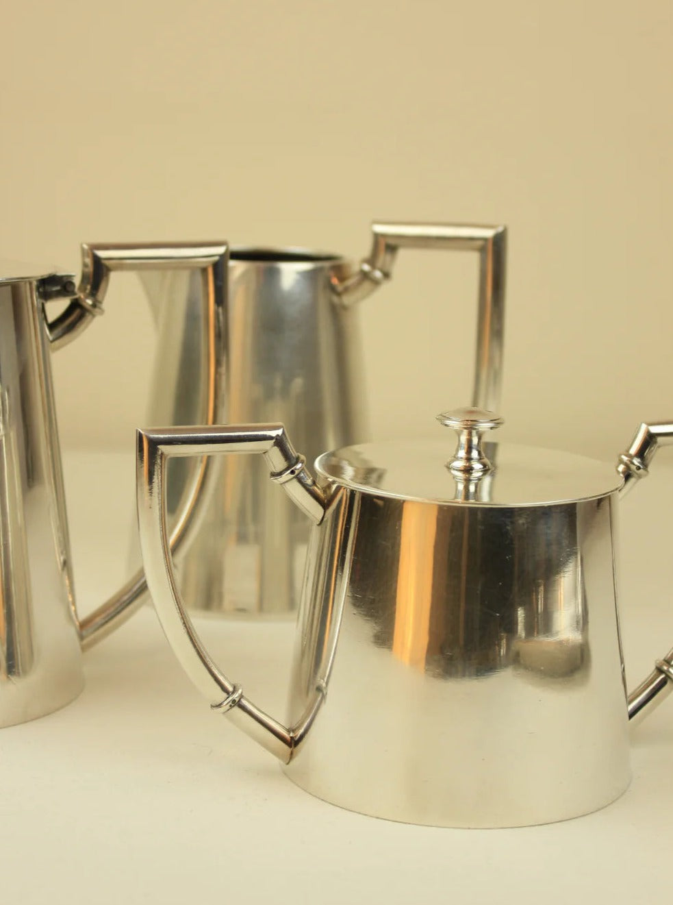 Two shiny silver Art Deco Tea/Coffee Services with curved handles on a beige background. The front teapot has a visible lid. Brand: Boga Avante Shop.