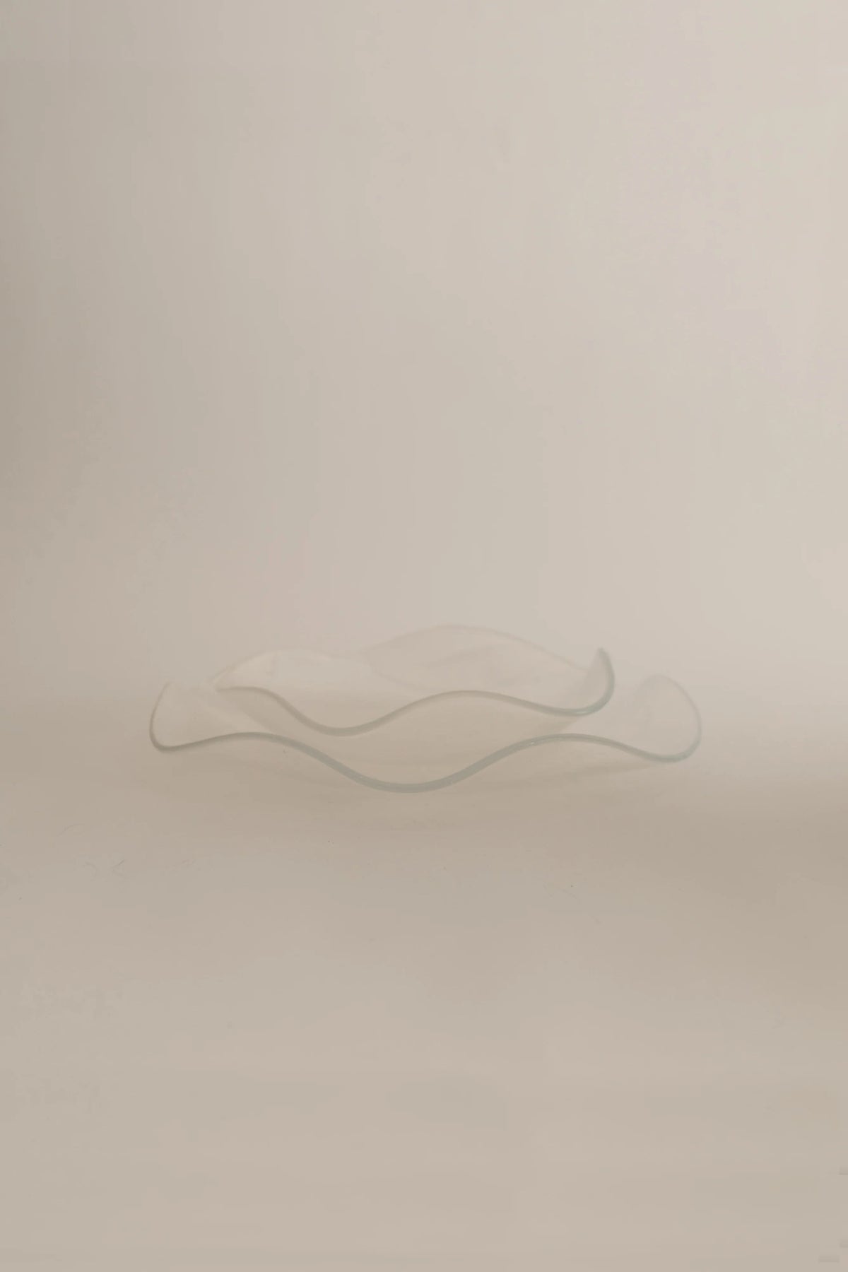 A translucent glass sculpture resembling a flowing abstract form, placed on a soft beige background, with a gentle gradient of light highlighting its curves. This hand-made creation embodies the meticulous intricacies of Sophie Lou Jacobsen's Petal Plate - Large.