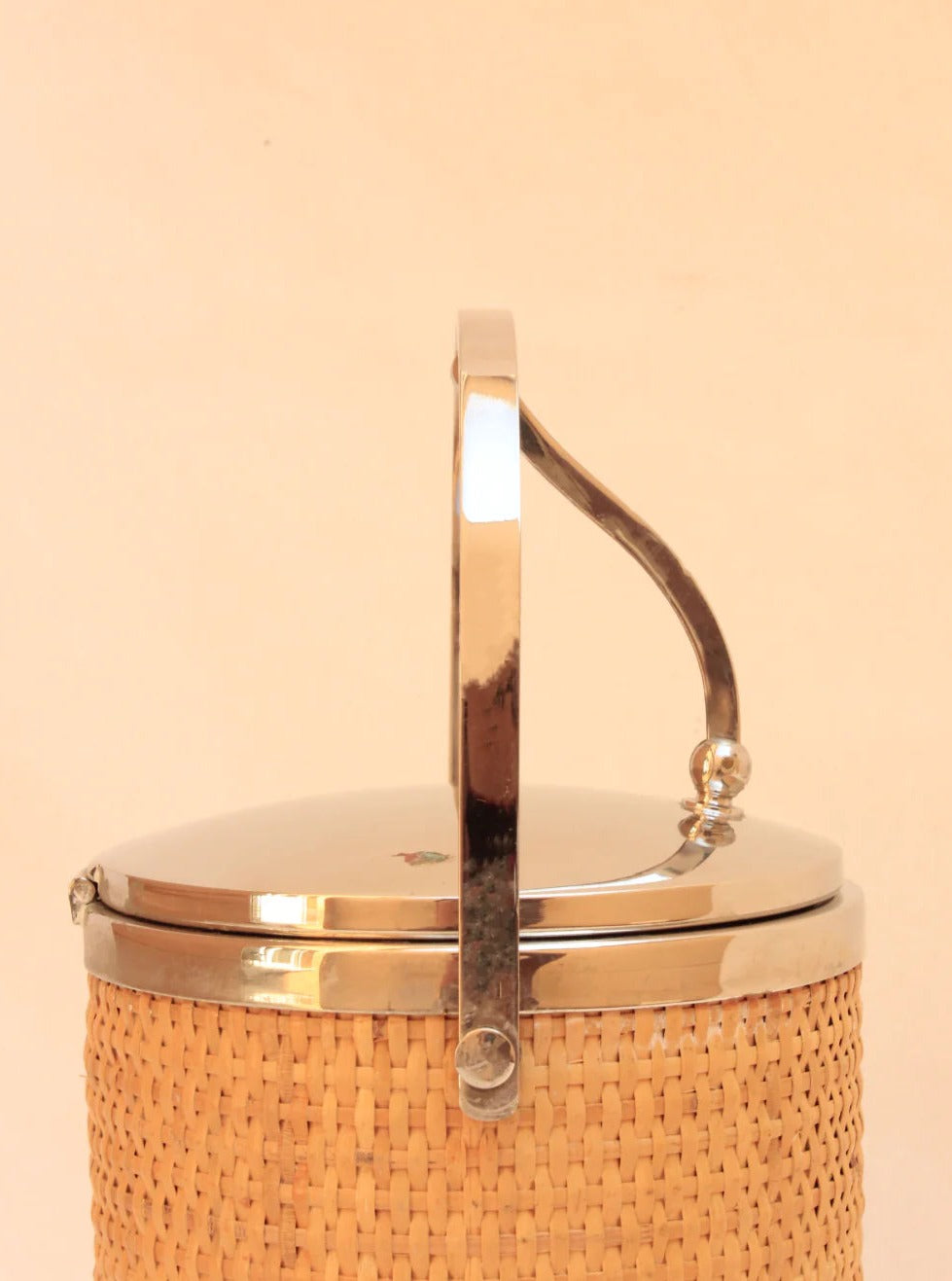 The Ratan Ice Bucket by Boga Avante Shop is a handcrafted cylindrical piece featuring a wicker texture with a circular metallic lid and handle, set against a beige background.
