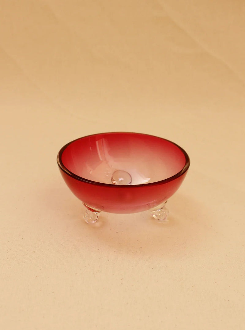 The Pink Murano Bowl from Boga Avante Shop is a small, round, red glass bowl that features a gradient effect transitioning to clear at the bottom. This handcrafted piece has a smooth surface and is supported by three clear glass feet, adding a vintage allure. The background showcases a neutral beige color.