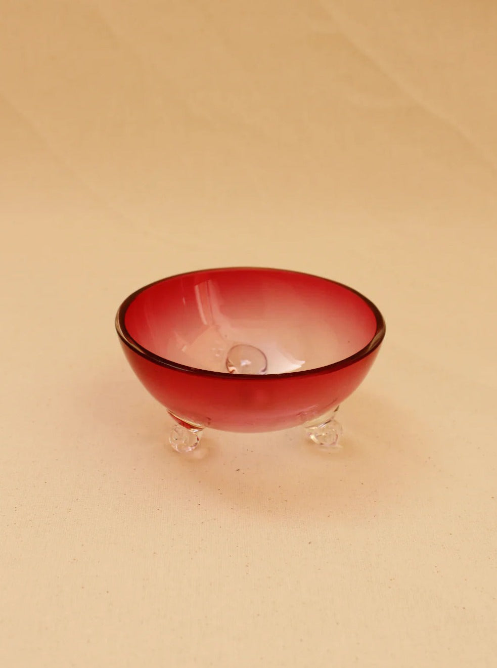 The Pink Murano Bowl from Boga Avante Shop is a small, round, red glass bowl that features a gradient effect transitioning to clear at the bottom. This handcrafted piece has a smooth surface and is supported by three clear glass feet, adding a vintage allure. The background showcases a neutral beige color.