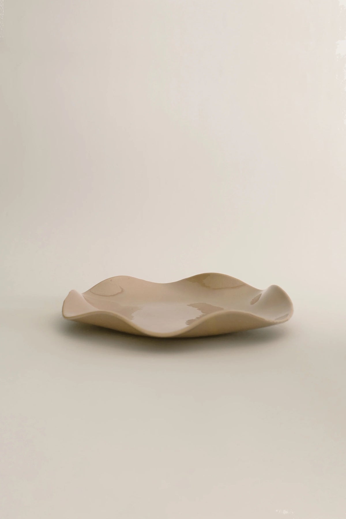 A Petal Plate - Large by Sophie Lou Jacobsen, with an irregular, wavy edge, set against a soft, neutral background. This artisanal plate exhibits a subtle reflective sheen with patches of light shadows.