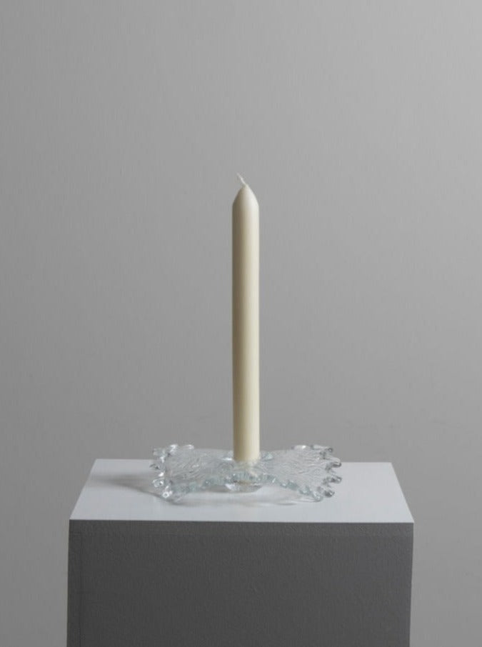 A single long white candle stands upright in the Uno Westerberg Candle Holder by Les Objets Amsterdam. The setup is positioned on a simple, light grey pedestal against a plain, light grey background. The scene is minimalistic and clean.