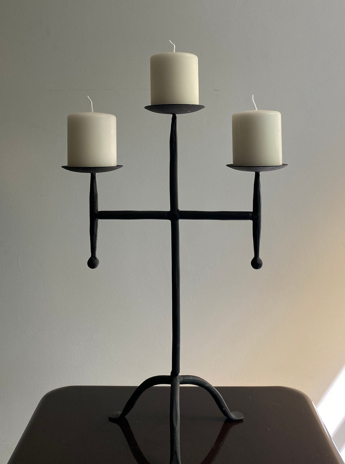 The Vintage Postmodern Iron Candle Holder by Les Objets Amsterdam is a sculptural black metal piece featuring three unlit white pillar candles. Crafted in the shape of a cross, it stands elegantly on a dark surface against a light wall, adding a luxurious touch to any interior.
