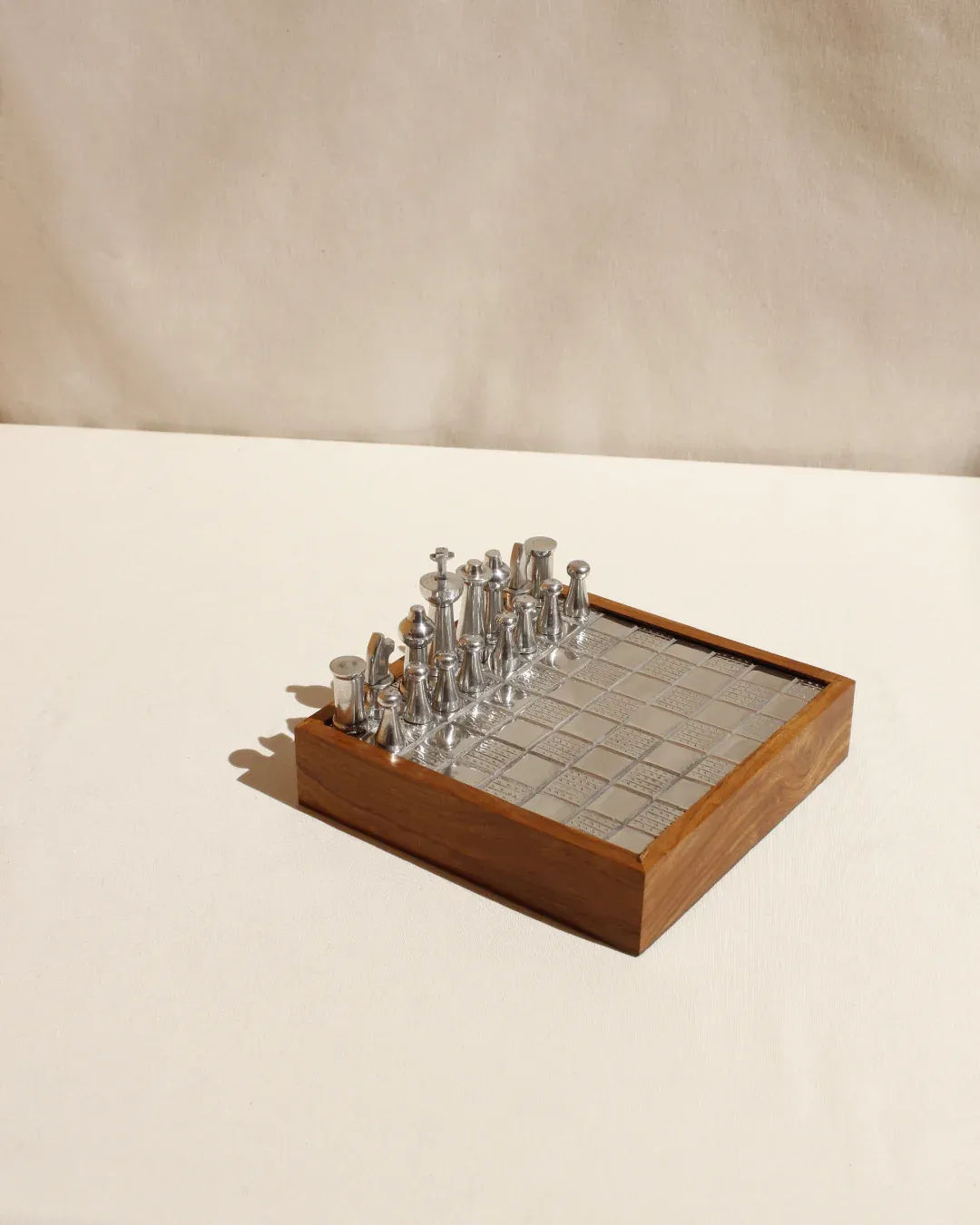 The Wood and Metal Chessboard from Boga Avante Shop features sculptural silver pieces on a wooden board with a metallic grid. Set against a beige surface, this minimalist design highlights one side of the board for an understated contemporary look.