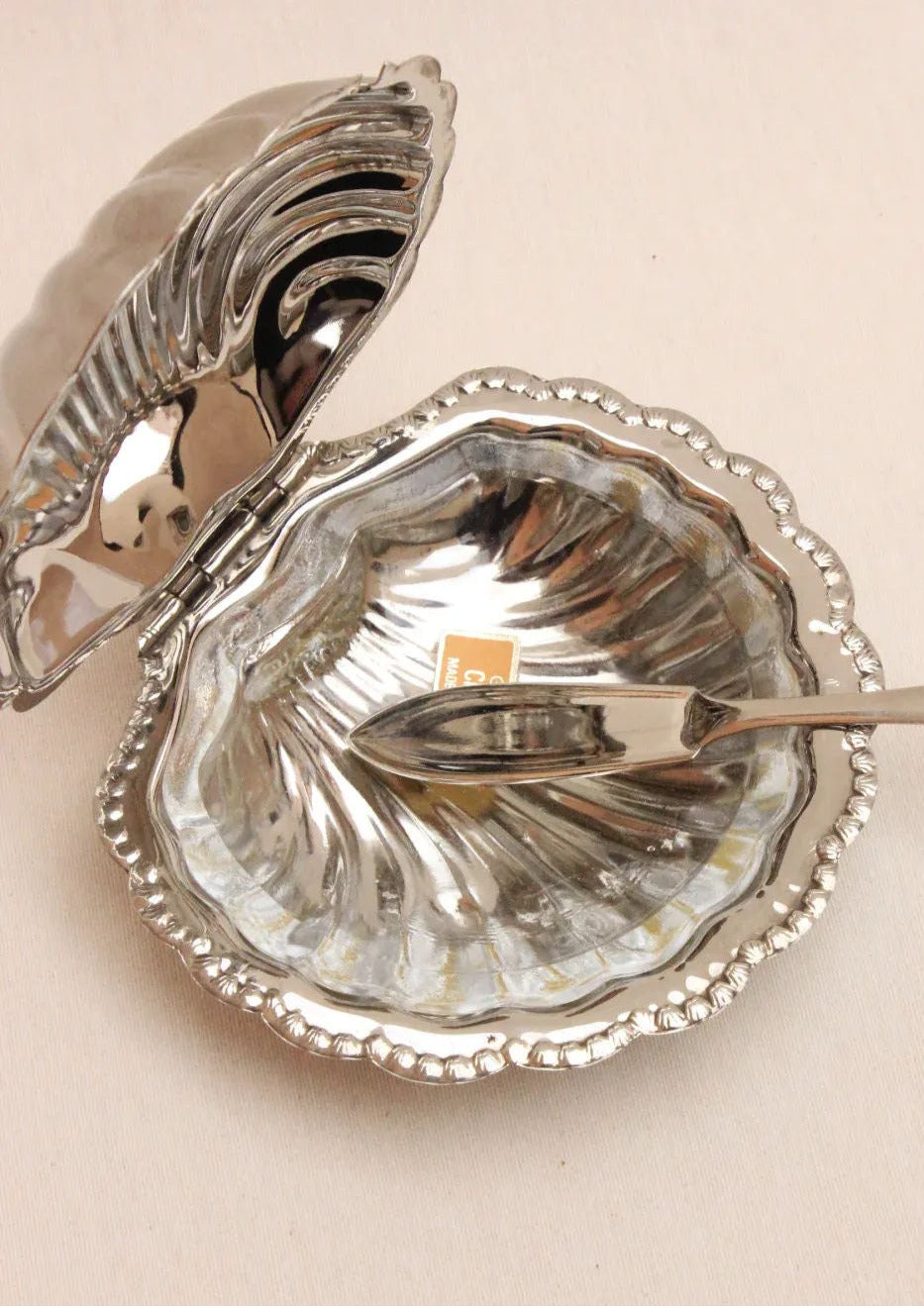 The Shell Shaped Butter Dish by Boga Avante Shop is an elegant piece with a textured interior and ornate wavy edges. Handcrafted to epitomize luxury, it includes a small rectangular tag and matching silver spoon, beautifully resting on a light, neutral surface.