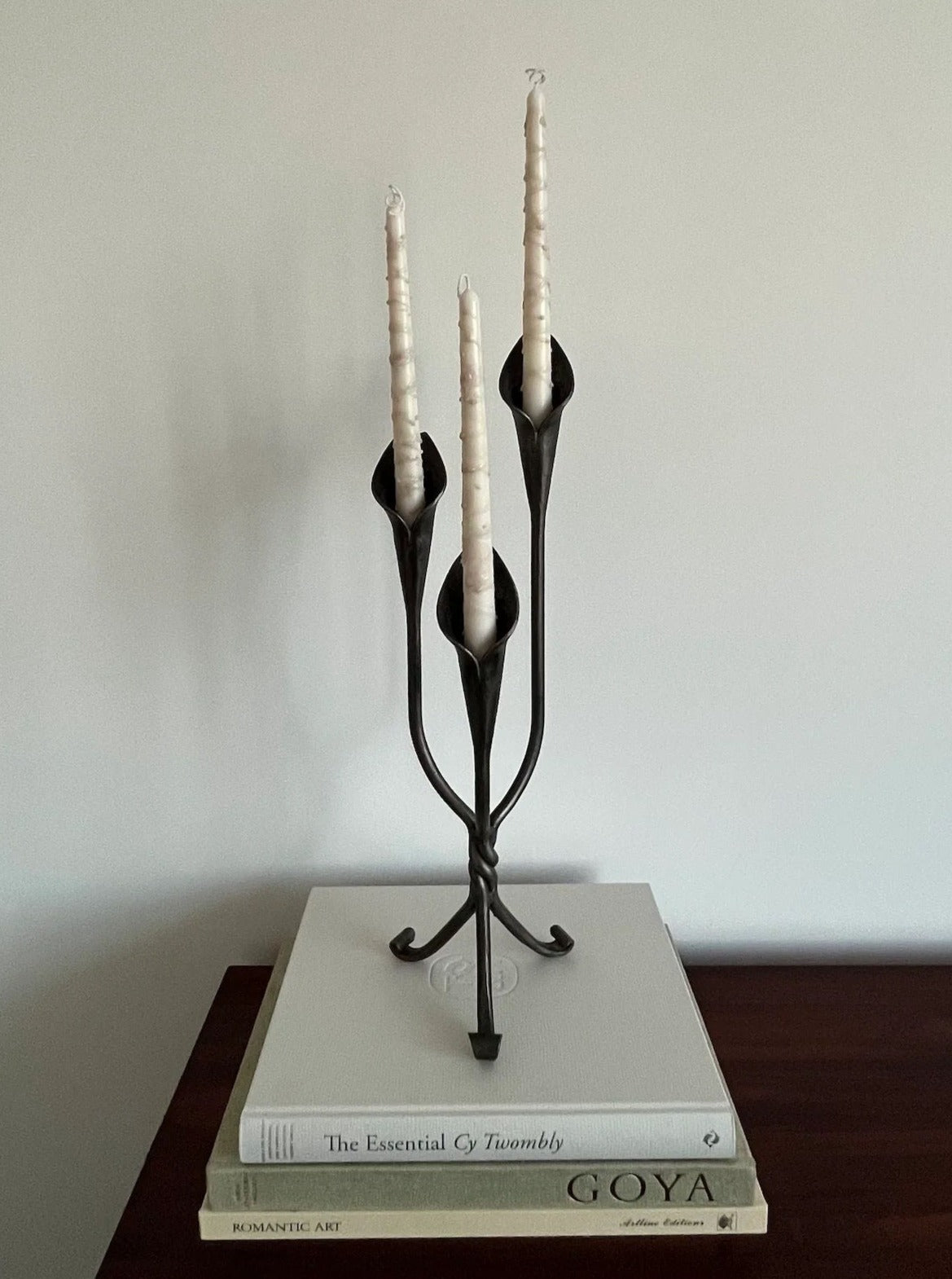 The Modernist Large Steel Tulip Candle Holder by Aesthete Label, topped with three spiral candles, sits atop stacked art books on a dark wood surface against a plain white wall for a touch of contemporary décor.