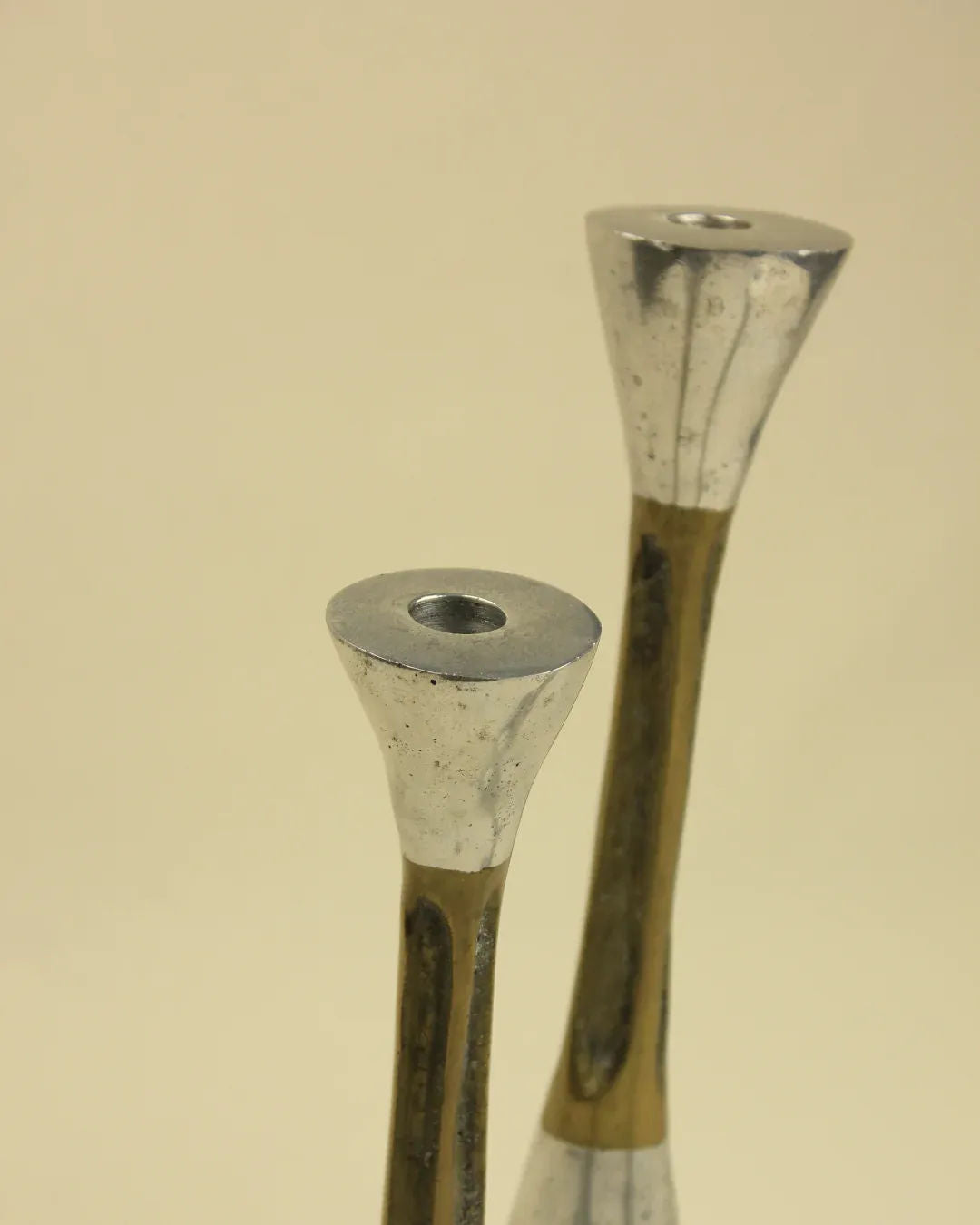 A set of three sleek metal candle holders, named the Trio of Brutalist Candleholders by Boga Avante Shop, are showcased against a neutral background. With tapered bases and wider tops, these holders evoke a retro 1980s charm with their modern design accented in both silver and bronze tones. Each holder features a small hole at the top for securing candles, making them perfect for adding flair to your home decor.