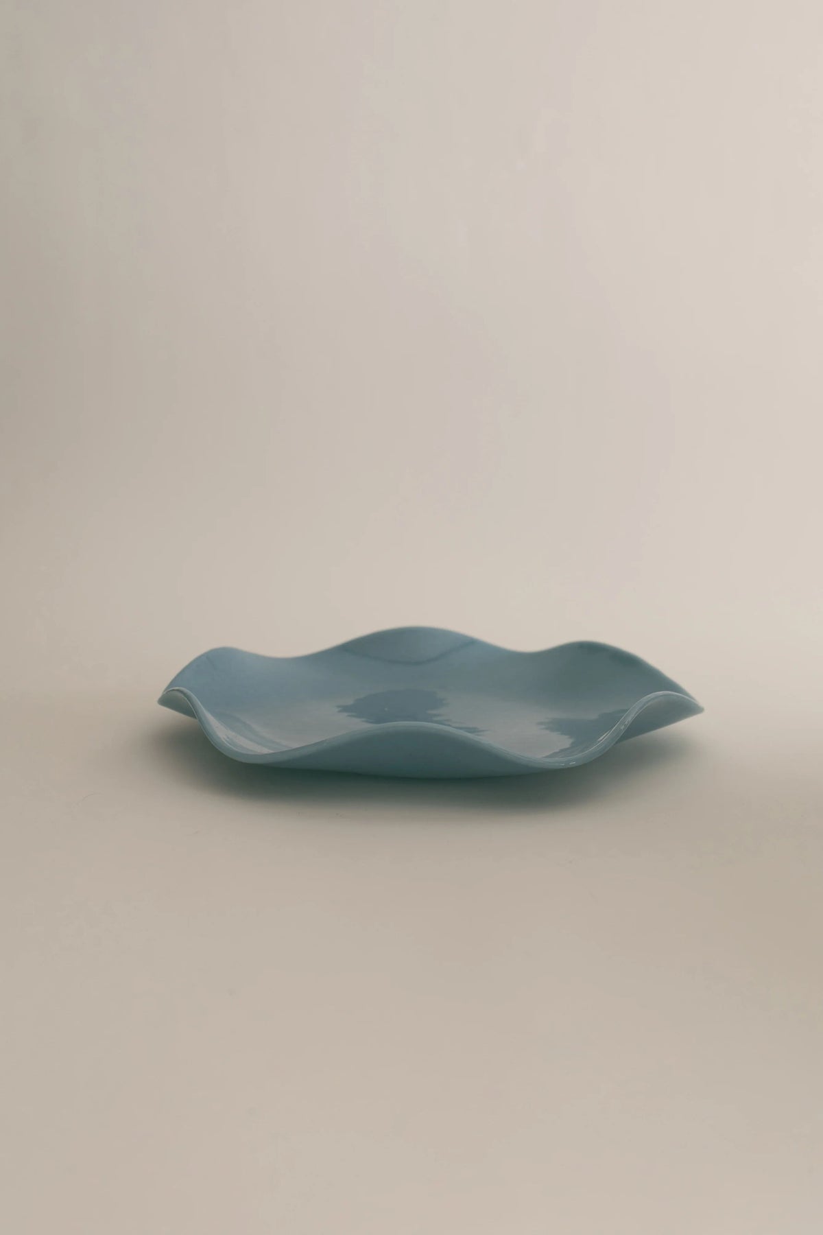 A minimalist blue ceramic Petal Plate - Large with a wavy design, hand-made through an artisanal production process, placed against a neutral beige background by Sophie Lou Jacobsen.