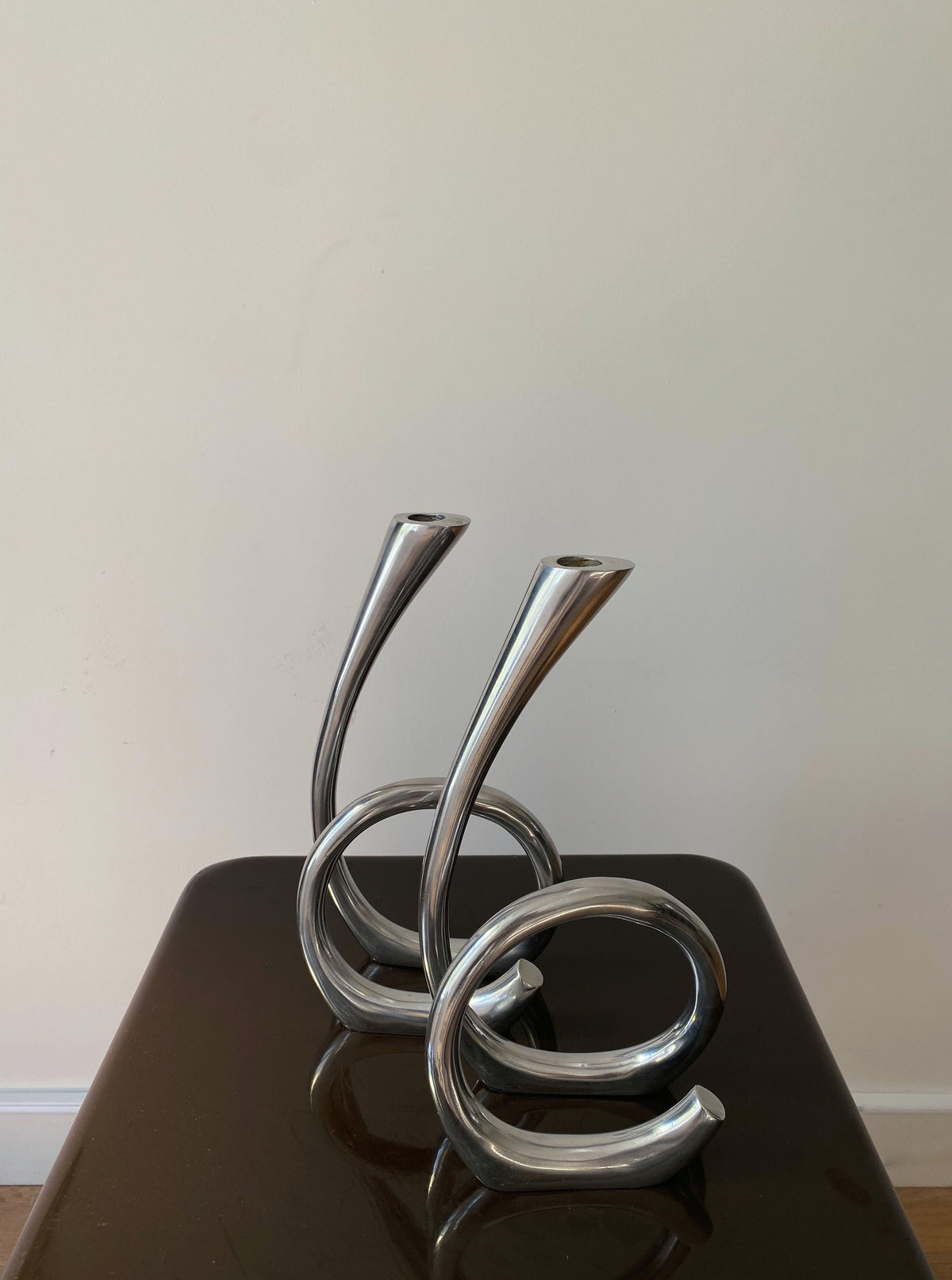 Two intertwined, twisted Vintage Curly RVS Candle Holders decorative sculptures from Les Objets Amsterdam, placed on a dark wooden surface against a plain light beige wall.
