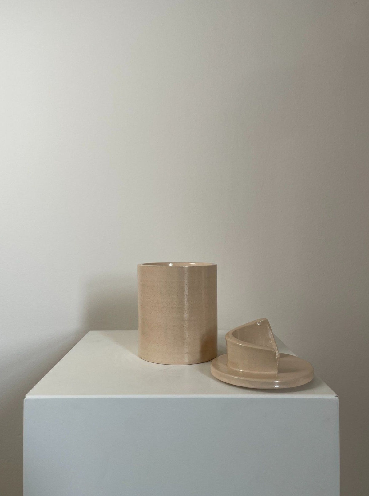 A minimalist display featuring a beige ceramic cup with its saucer placed slightly askew, and the Sculptural Scented Candle No. 11 from 2222Studio, all set atop a white pedestal against a pale grey background