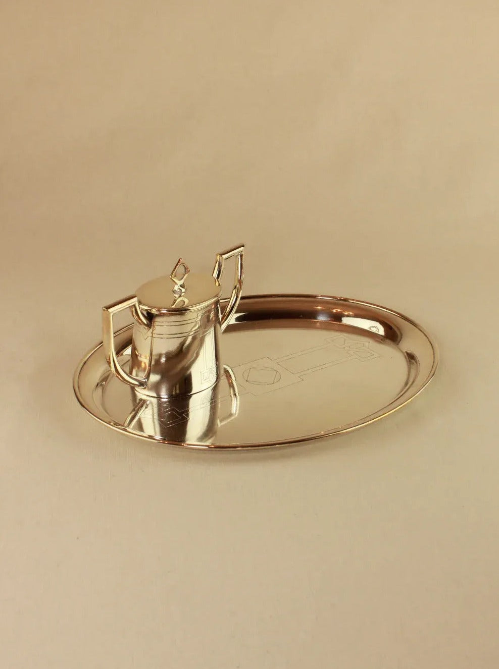 A Boga Avante Shop Art Deco Tea Set with a silver teapot, lid, and handle, displayed on a matching oval silver tray in the Art Deco style, set against a neutral beige background.