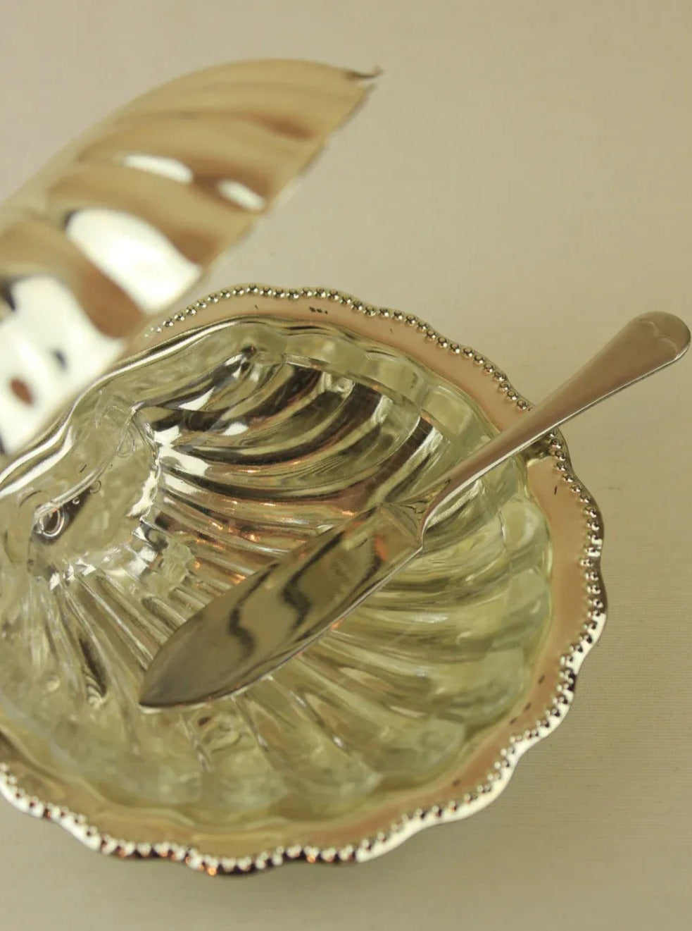 The Boga Avante Shop's Shell Shaped Butter Dish features a polished silver finish with a matching lid, slightly ajar to reveal its inner compartment. A sleek, decorative butter knife with a smooth handle rests inside the dish, positioned diagonally. The dish boasts a reflective, shiny surface that beautifully complements the intricately designed knife.
