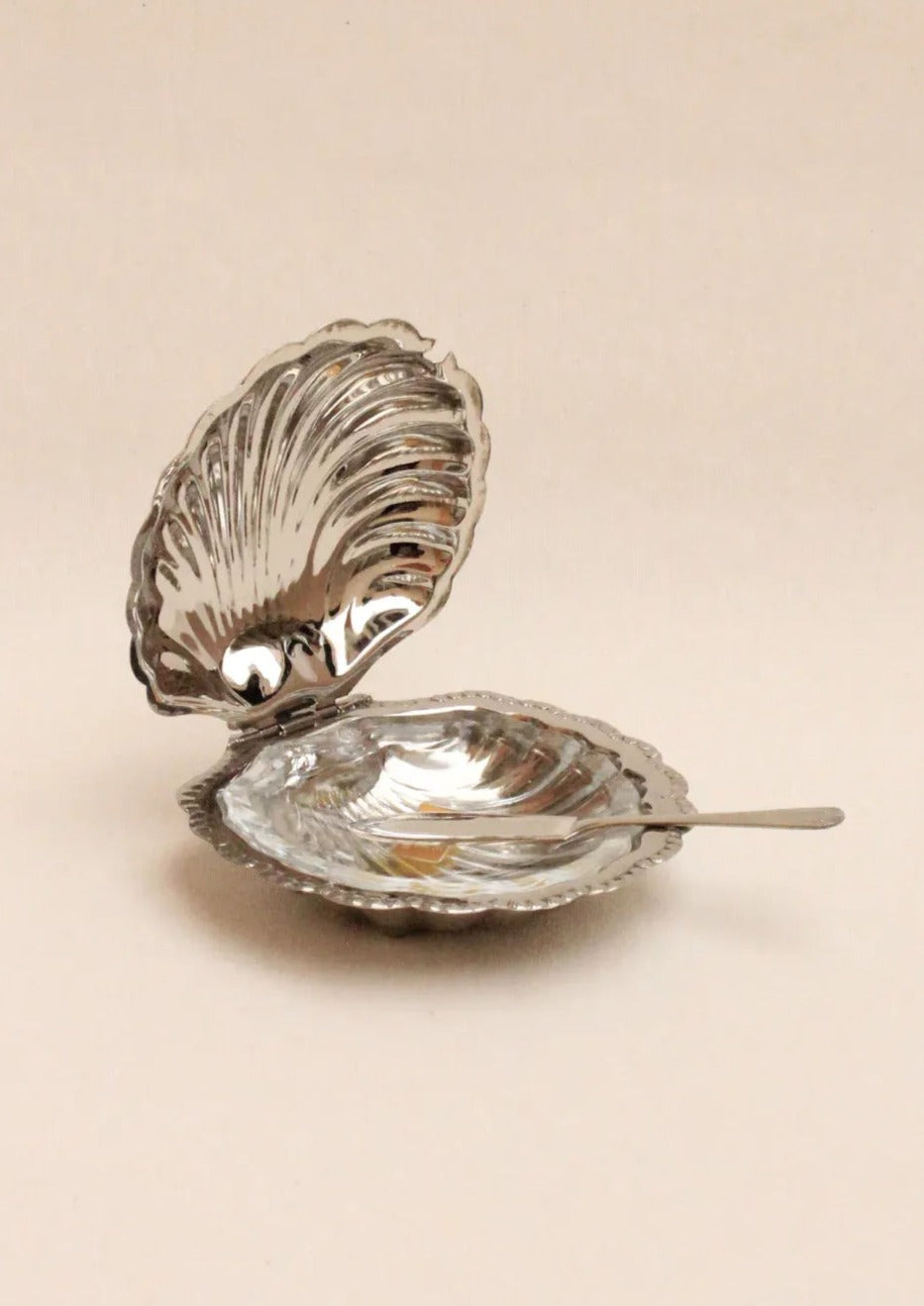 The Boga Avante Shop's Shell Shaped Butter Dish features a metallic, sculptural design with detailed ridges and a glossy finish. The open lid reveals a matching spoon, epitomizing minimalist aesthetics against a neutral background.