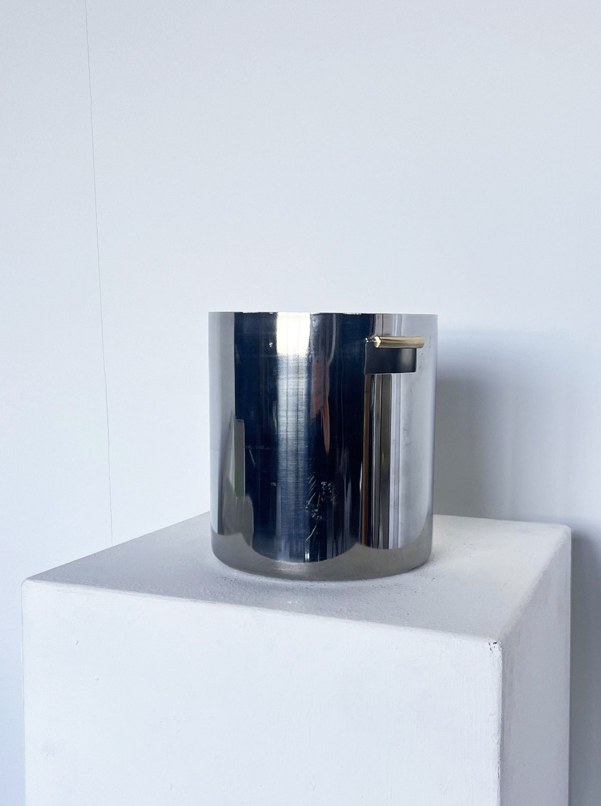 A shiny silver cylindrical Brutalist ice bucket with a brass detail handle, placed on a white pedestal against a white wall by Porter Studio.