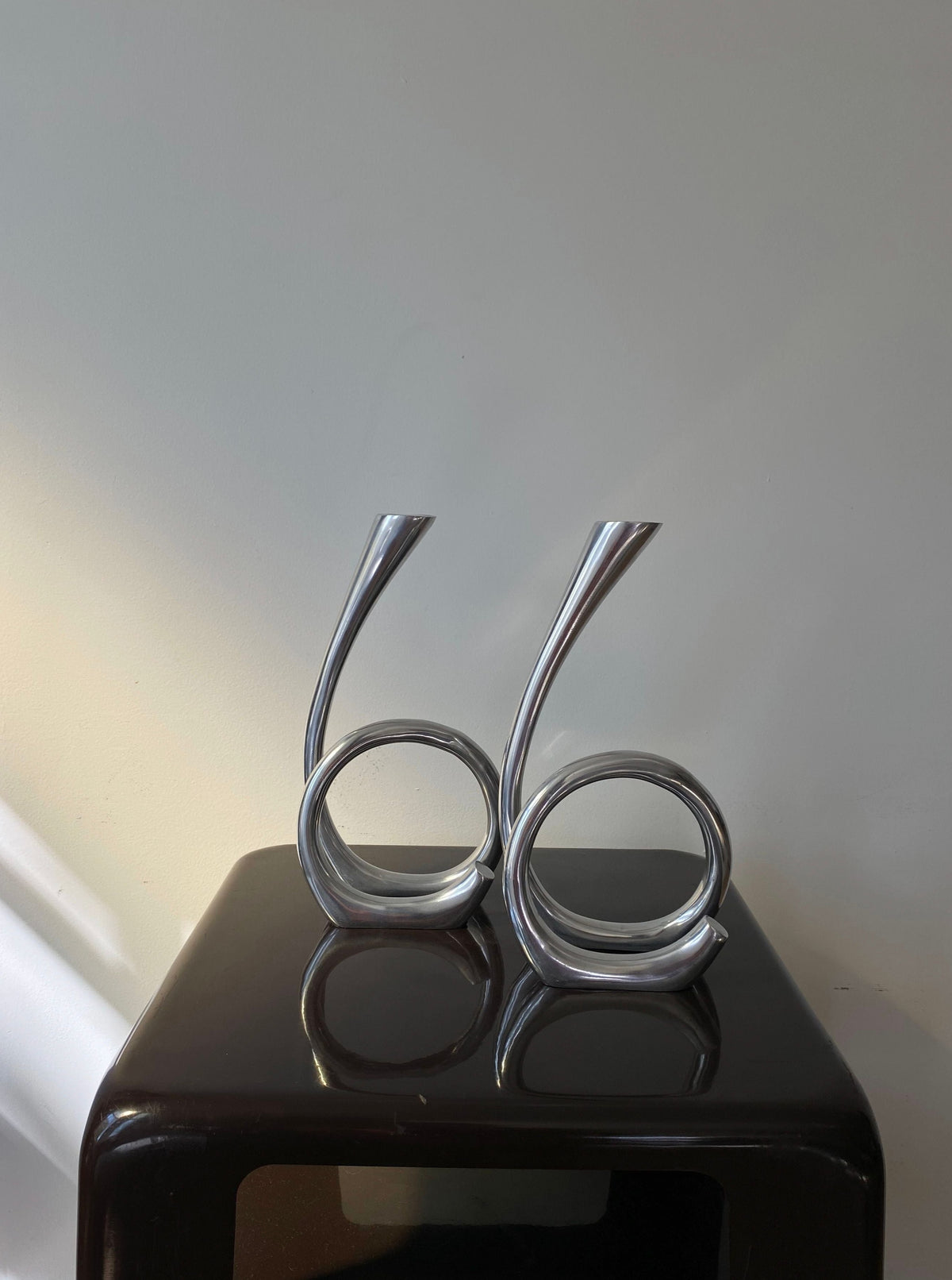 Two sleek, silver, Vintage Curly RVS Candle Holders standing upright on a glossy black surface against a light background. The curls emulate abstract engagements of infinity symbols by Les Objets Amsterdam.