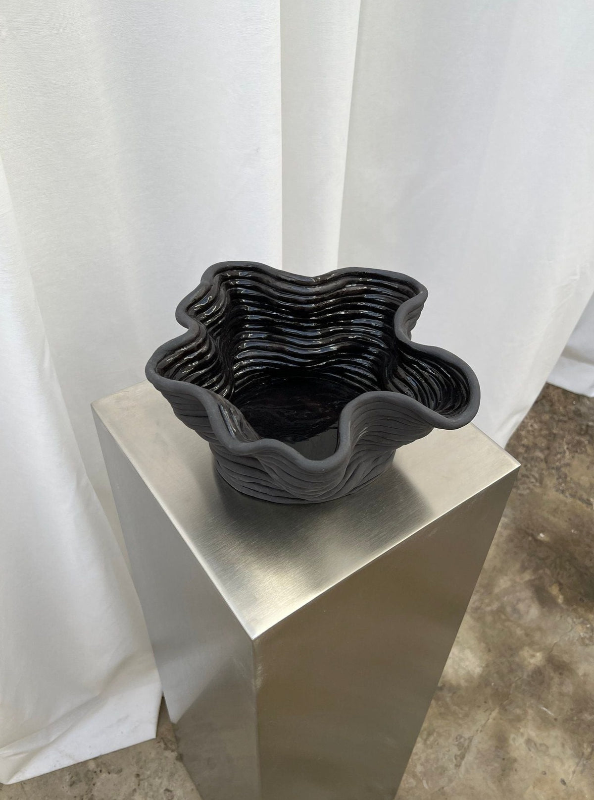 The Wobbly Bowl Black by 2222Studio, a sculptural ceramic piece with a wavy rim and layered texture, rests on a metallic pedestal against white curtains and a stone floor, highlighting its minimalist contemporary design.