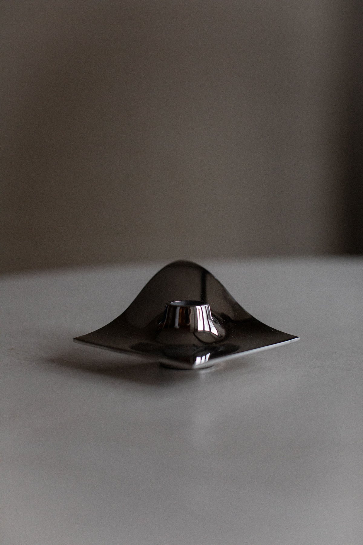 A sleek, metallic Out For Lunch 70's Chrome candle holder in a minimalist style sits on a gray surface, displaying a unique undulating design and reflective surface.