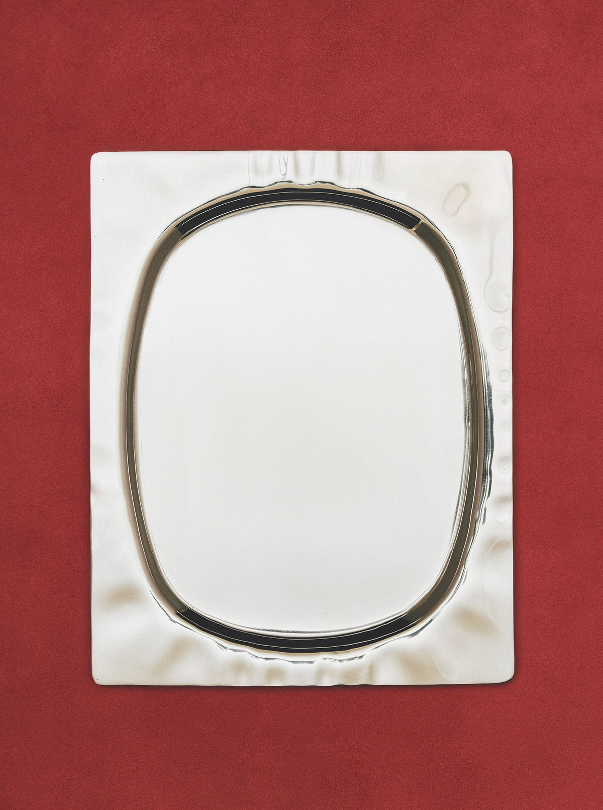 A square silver tray with a polished finish and a reflective, oval-shaped center placed on a deep red background with a textured surface. (Product Name: 104-1 by Ananas Ananas)