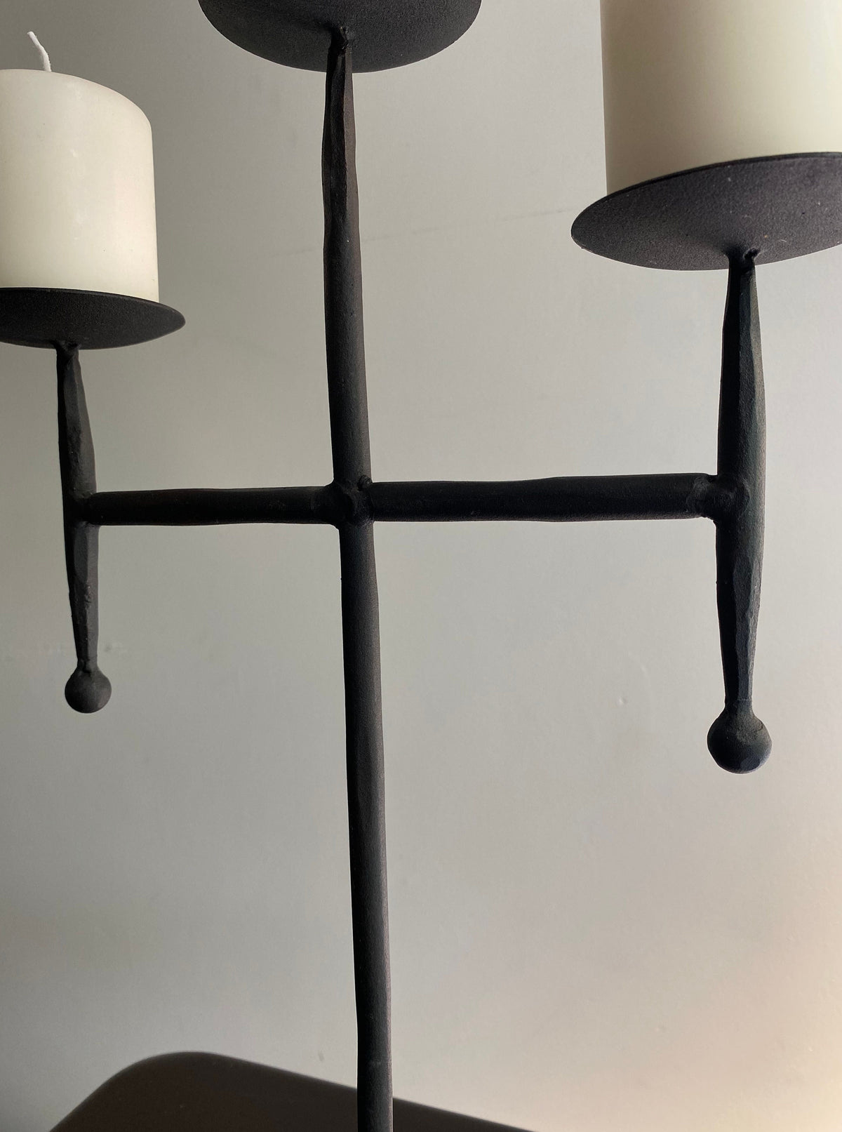 The Vintage Postmodern Iron Candle Holder by Les Objets Amsterdam cradles two unlit cream pillar candles against a plain, light wall. Its sculptural, minimalist design adds elegance to any luxury interior.