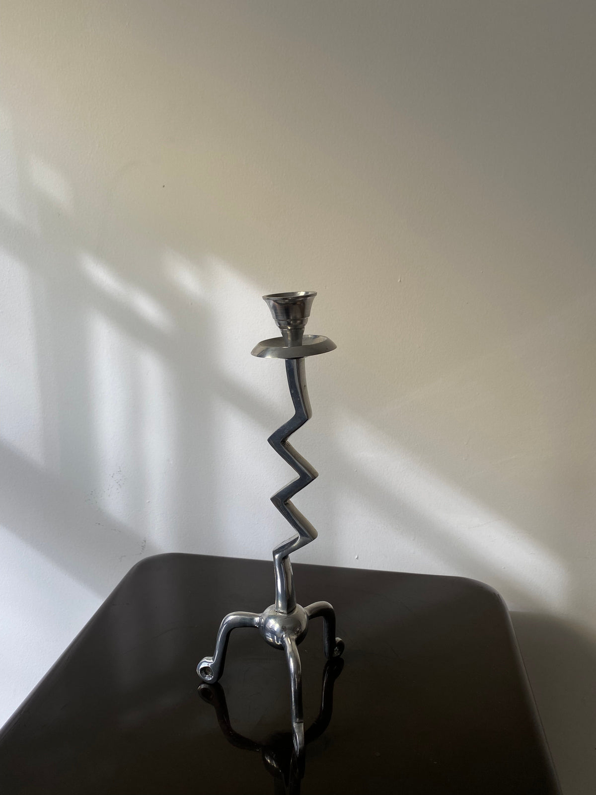 The Vintage Zigzag Candle Holder by Les Objets Amsterdam, handcrafted in silver with a zigzag stem and three sculptural legs, elegantly sits on a black surface, casting soft shadows on the white wall.