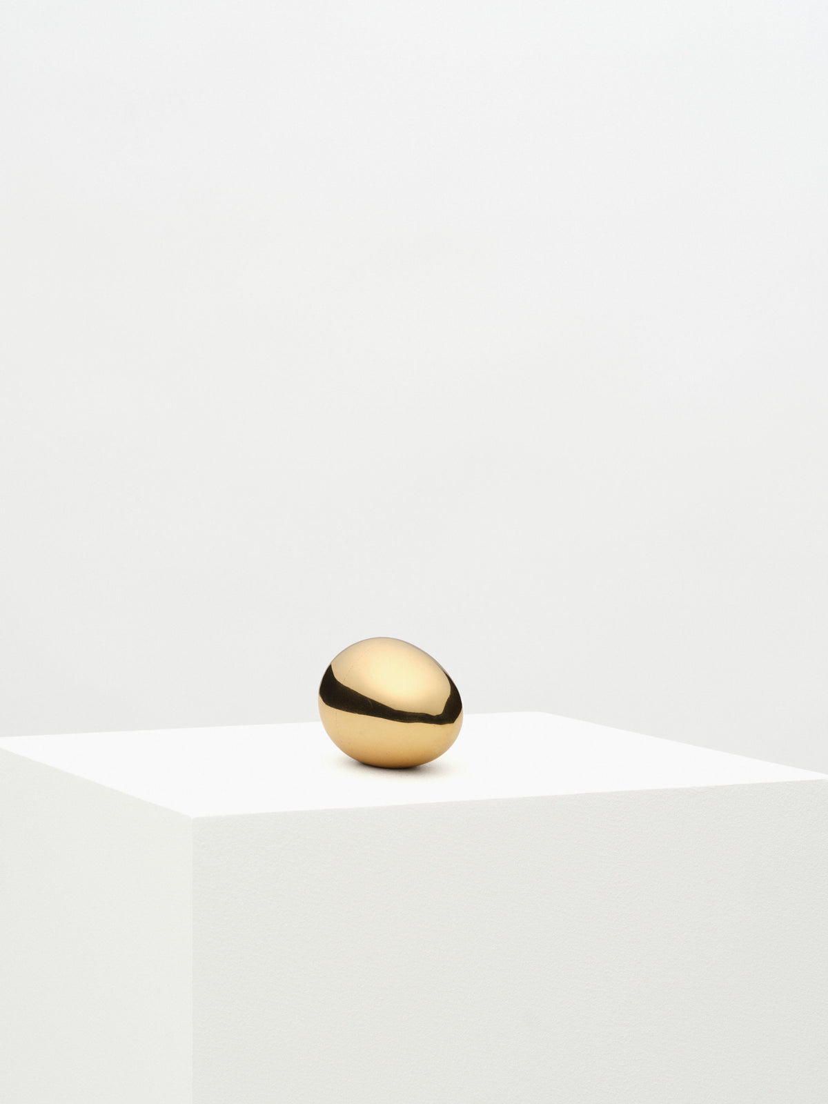 A simple brass Fourth St egg paperweight rests centered on a white pedestal against a plain, white background, portraying a minimalist and elegant visual.