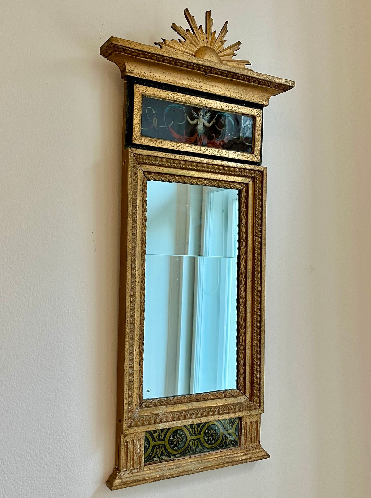 An Médecine antique gilded mirror framed in ornate gold with intricate detailing and decorations at the top, including a sunburst design. The reflection shows a glimpse of curtains.