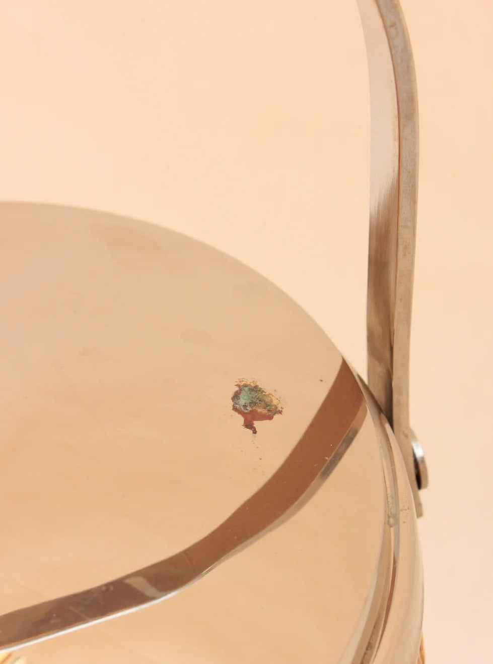 A close-up of the Boga Avante Shop's Ratan Ice Bucket reveals its sculptural metallic design with a curved handle, highlighting a small rust spot. The object stands out against a soft peach background, accentuating its contemporary aesthetic.
