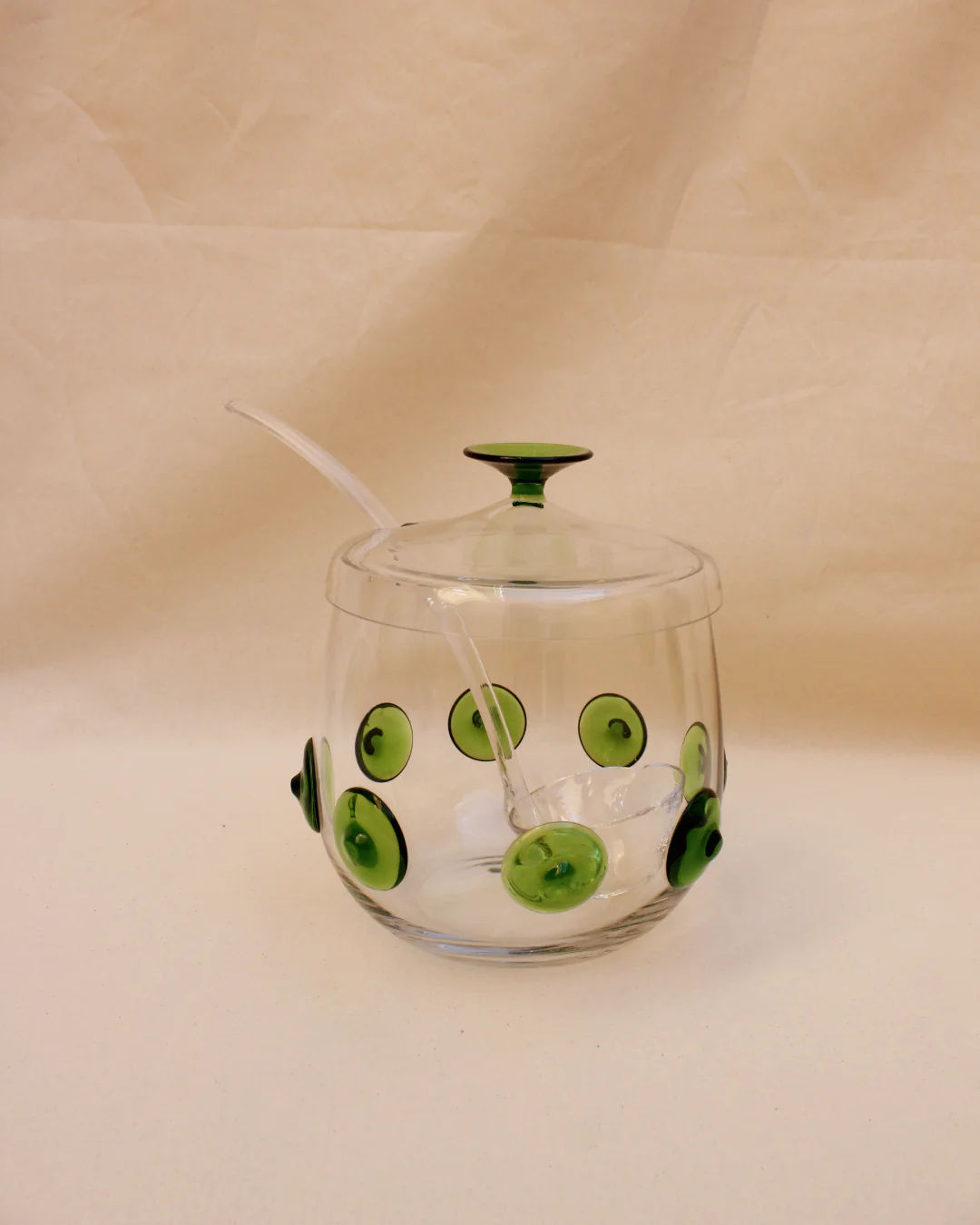 A clear glass Murano Punch Bowl with green circular and oval patterns, featuring a matching green lid and a transparent glass stirring spoon, displayed against a crumpled beige backdrop. This handcrafted piece serves as an ideal centerpiece.