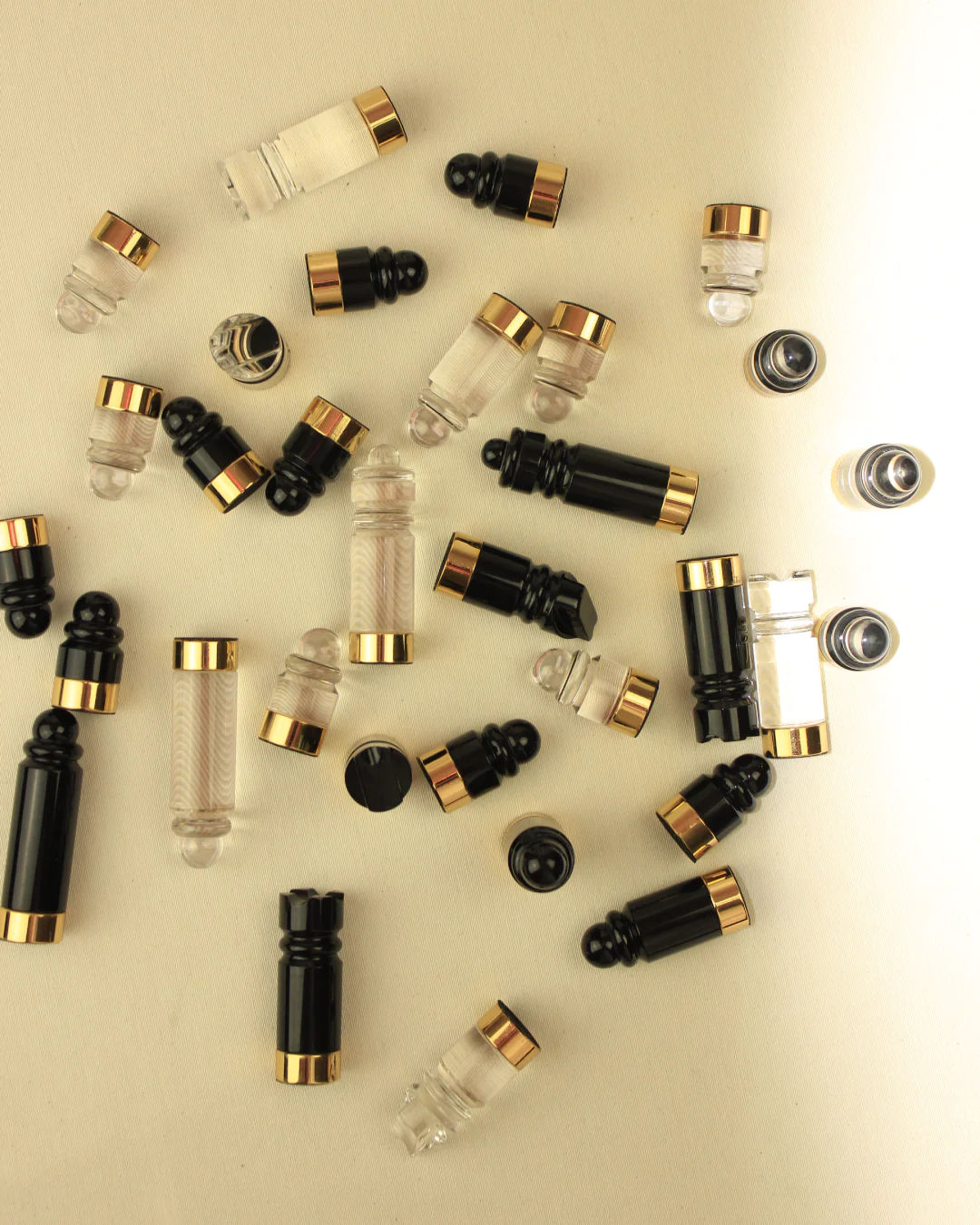Various small cosmetic bottles with black and gold caps, featuring brass accents, scattered on a light beige background. Some caps are separated from the Boga Avante Shop Chess & Checker Board.