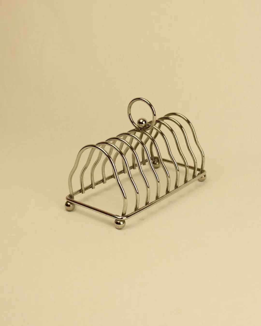 A silver stainless steel Art Deco Toast Holder from Boga Avante Shop with four curved slots on a plain beige background. The holder has a circular handle on top and ball feet.