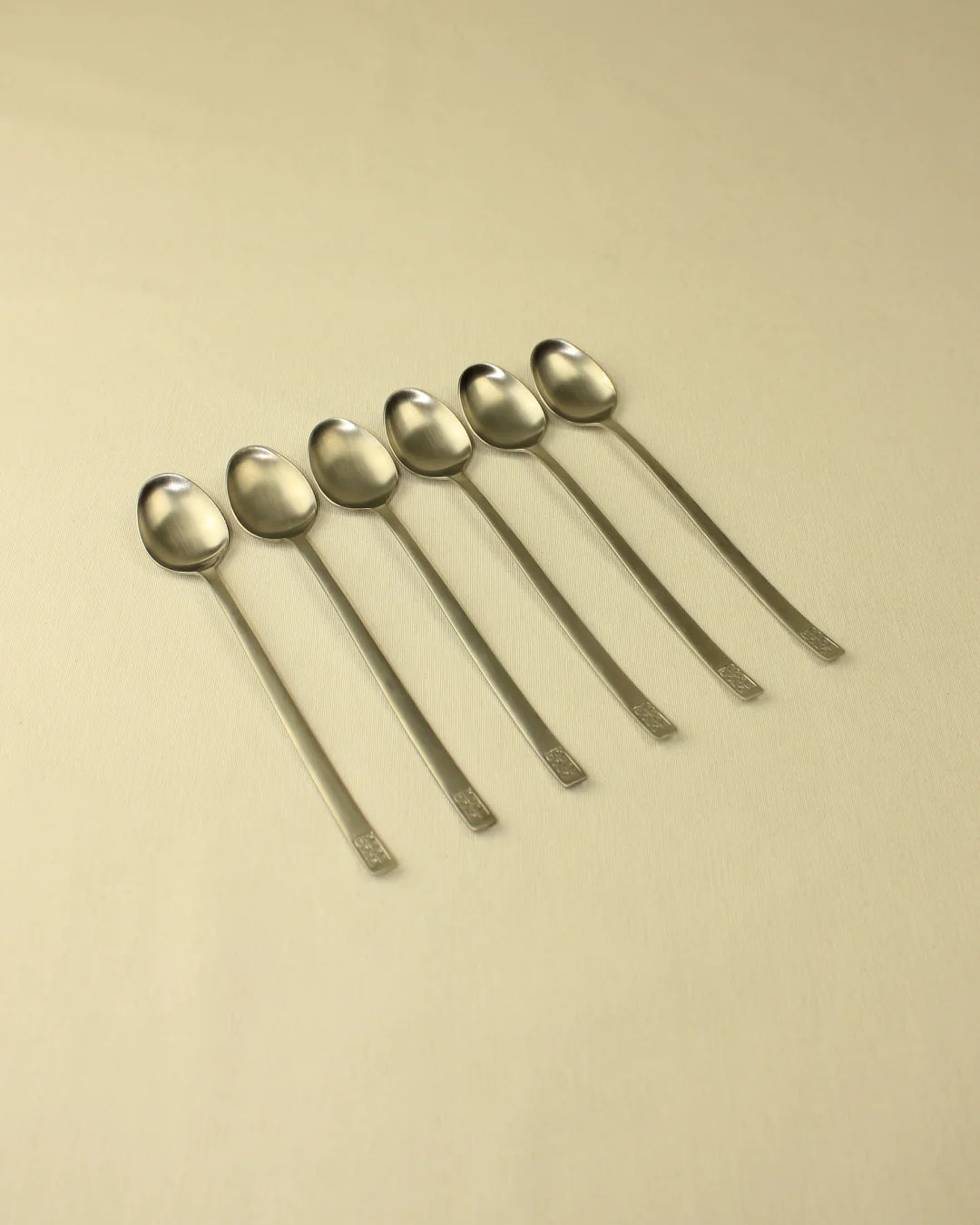 Six Boga Avante Shop 70s Style Spoons arranged in a line on a light beige surface, with their handles pointing right and heads gradually tilting towards the surface.