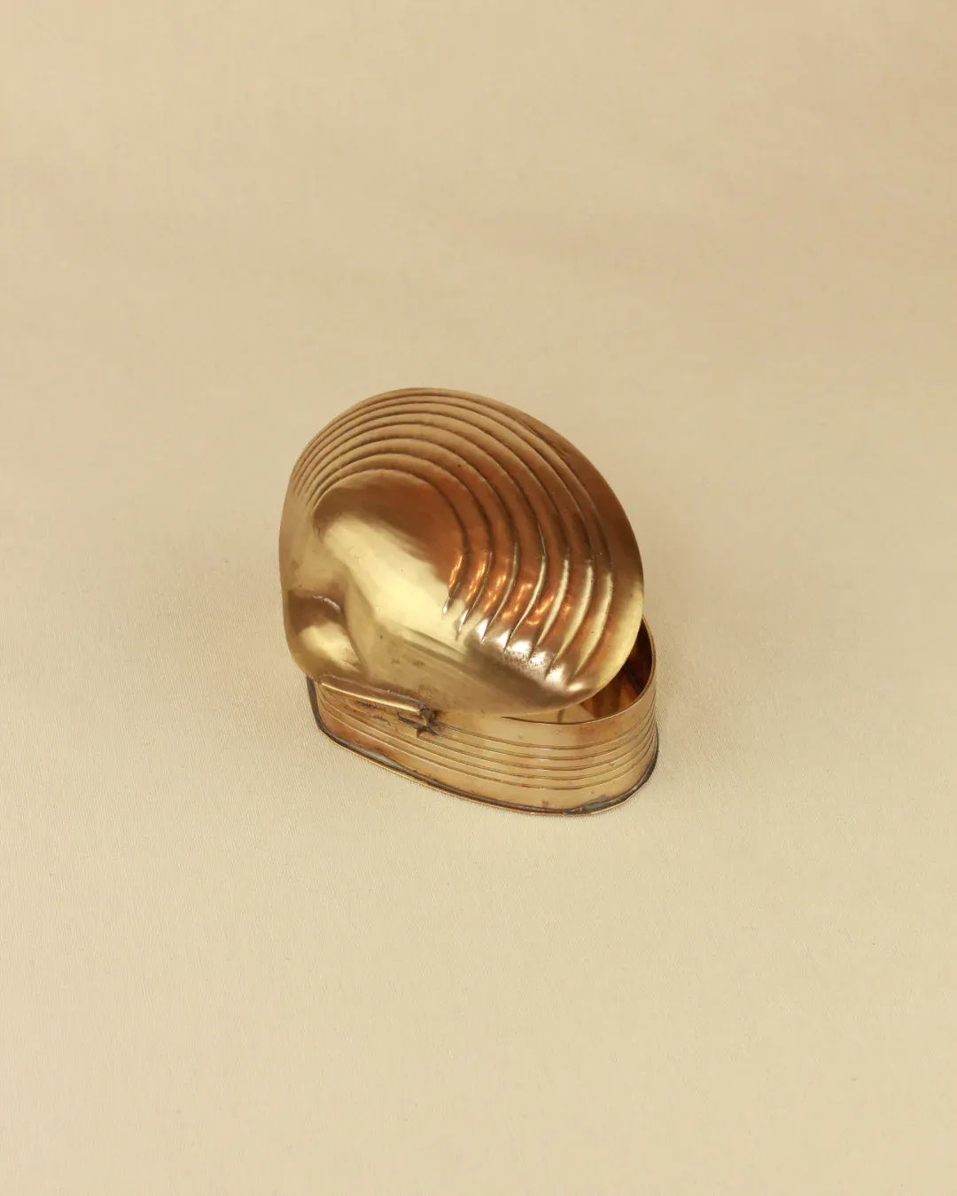 A Boga Avante Shop brass-hinged, seashell-shaped ornament on a light beige background, featuring detailed ridges and a smooth, shiny surface.