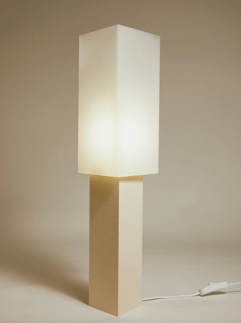 A modern, minimalist Japanese design Lampe ii table lamp with a glowing square top section mounted on a rectangular base, set against a soft beige background. The lamp casts a warm light and has a visible power cord from Argot Studio.