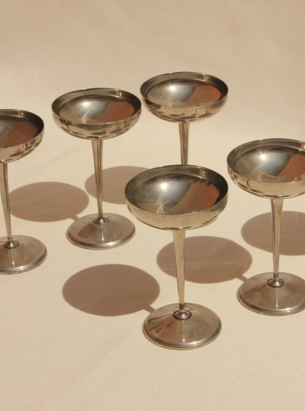 Five vintage Boga Avante Shop Alpaca Silver champagne coupes arranged on a sandy beige surface, showcasing their reflective bowls and elegant stems under soft lighting.
