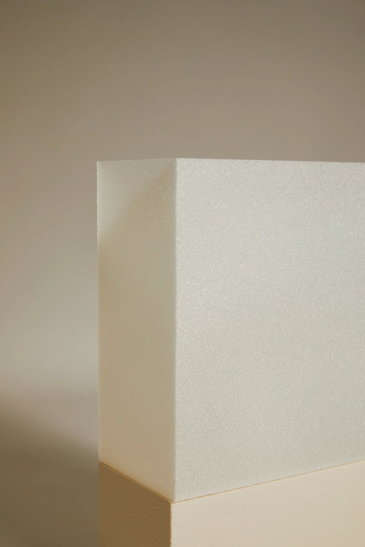 The Argot Studio's Lampe i is a sculptural, textured white block that stands upright on a reflective surface. The light casts subtle shadows, highlighting its geometric form against a beige gradient background, enhancing the contemporary and minimalist aesthetic of luxury interiors.