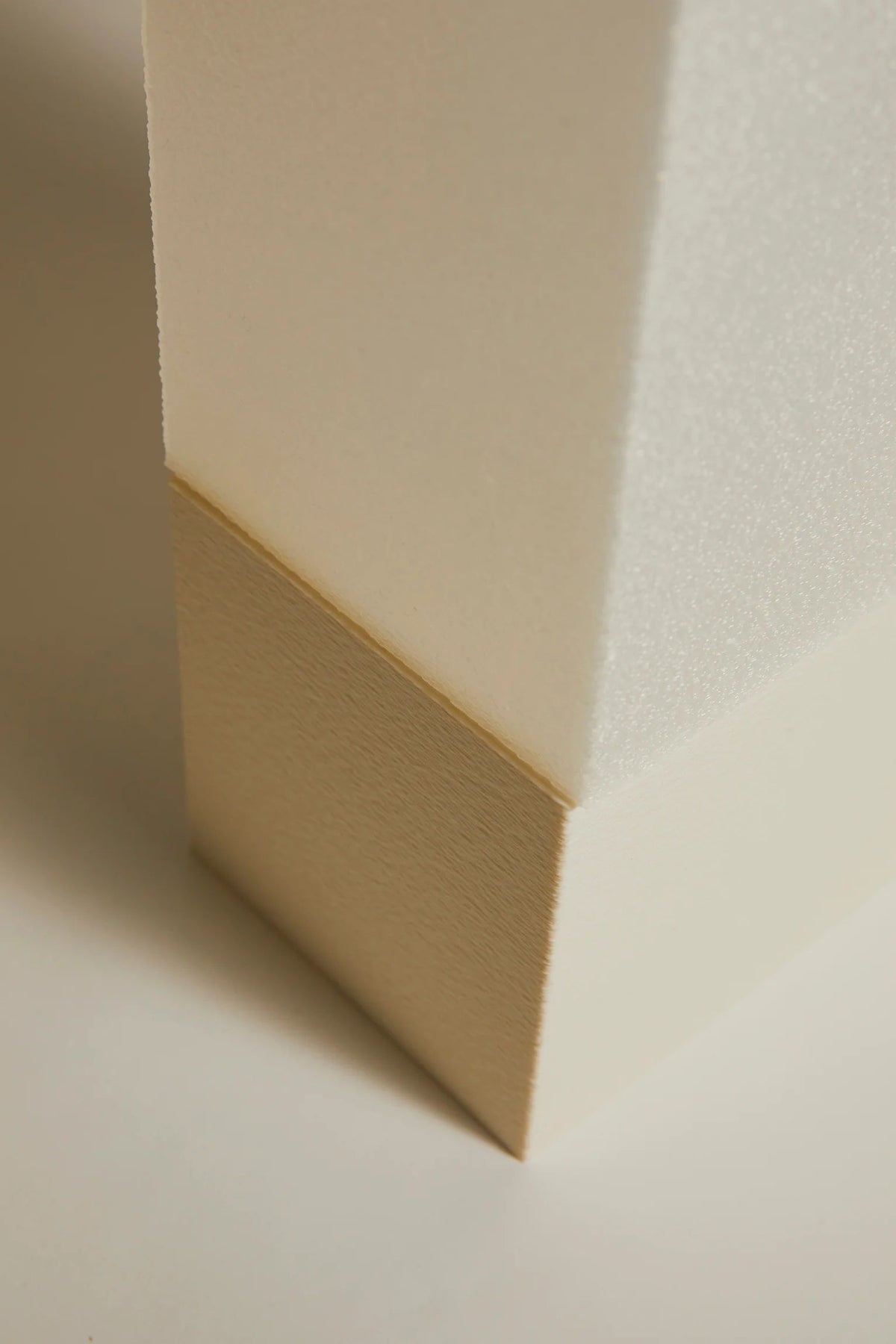 A close-up of Argot Studio's Lampe i reveals its sculptural, minimalist aesthetic: a textured rectangular object with a matte finish, featuring a smooth off-white upper section and beige lower section with subtle grain. Soft lighting highlights its form, casting gentle shadows.